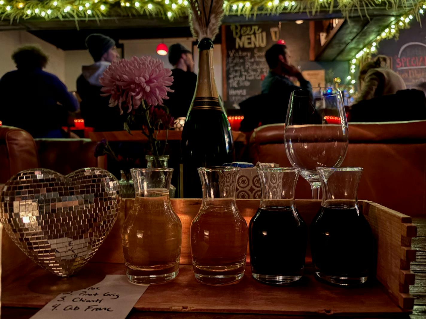 Four full tasting carafes with white and red wine next to a heart shaped disco ball on a wood table. The bar is visible in the background.