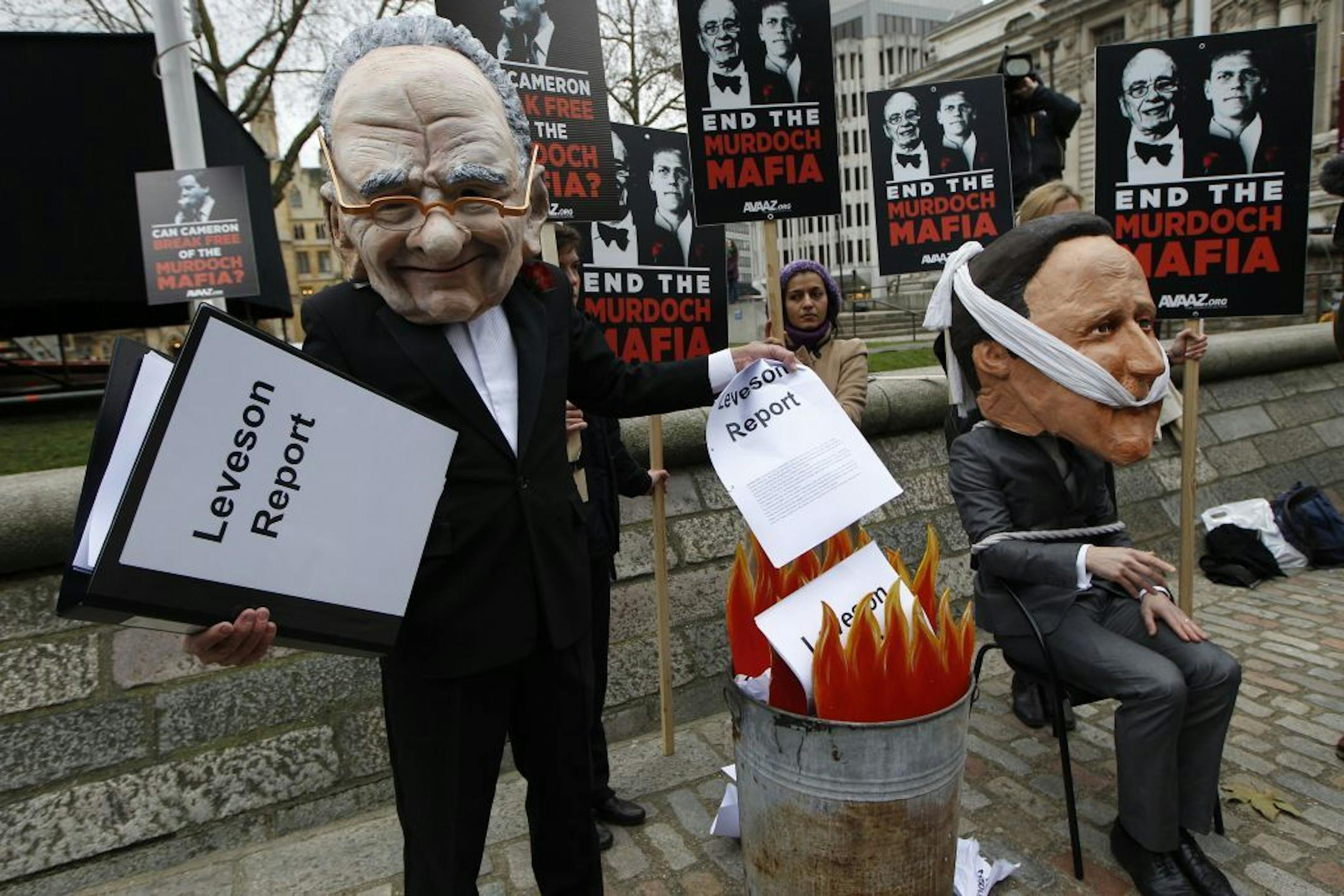 A campaigner wearing a giant mask depicting News Corporation's chairman Rupert Murdoch burns the Leveson report while another wearing a mask depicting British Prime Minister David Cameron, sits tied to a chair during a protest, calling on MPs to back reform legislation to stop any one media organisation developing a stranglehold over the British media, outside the Queen Elizabeth II Conference Centre in London where Lord Justice Brian Leveson is to release his report into the culture and practic