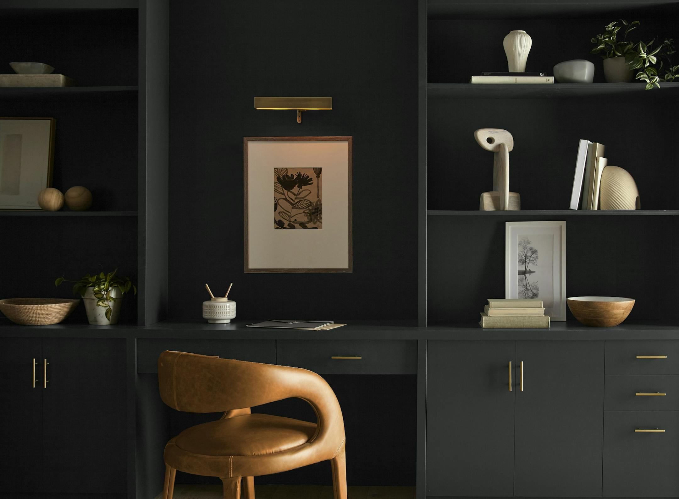 An office painted in soft black with dark gray built-in bookcases with gold handles and a tan leather chair.