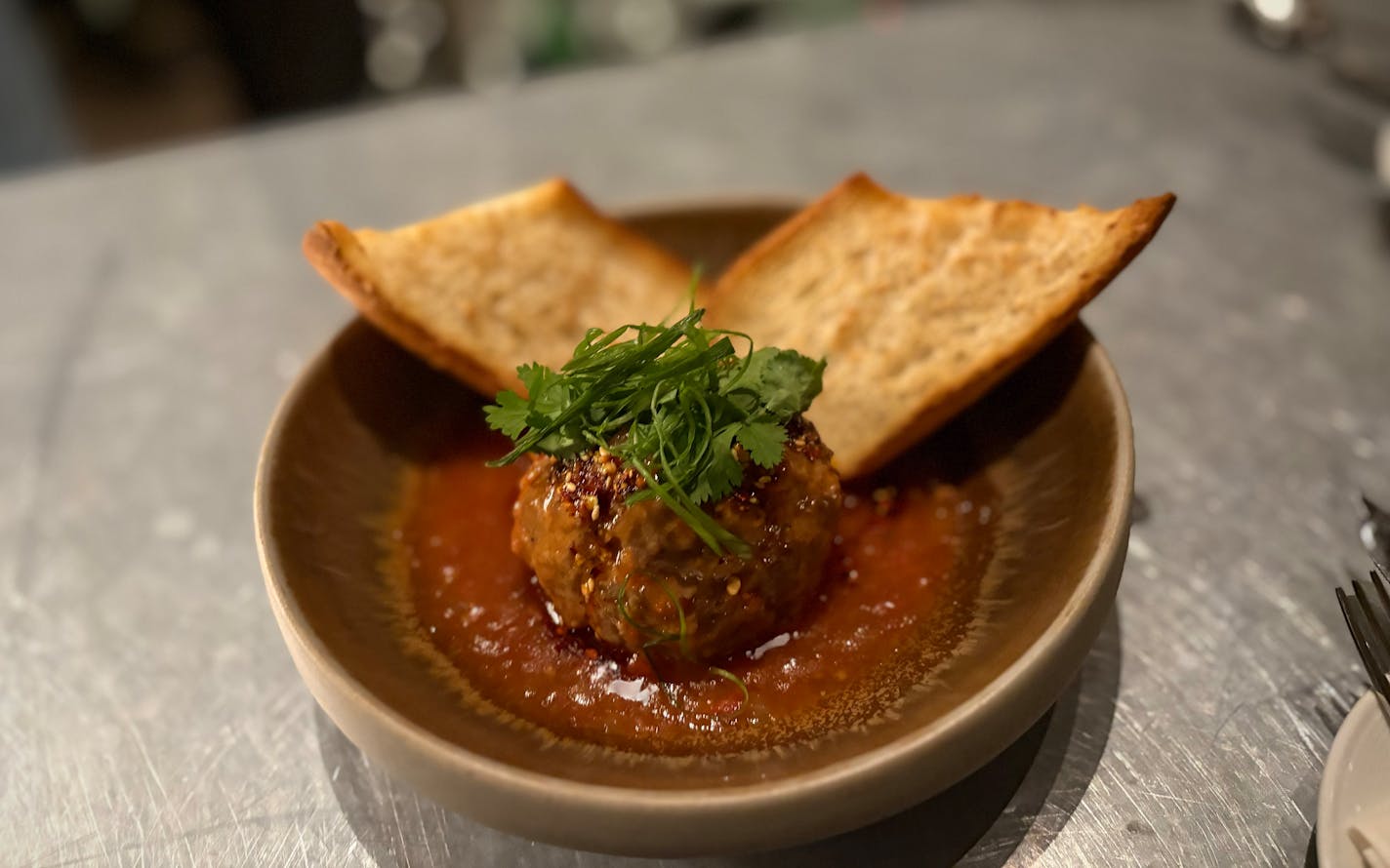 A large meatball topped with cilantro and chives is in a shallow bowl holding a pool of sauce. It's served with two slices of crusty Italian bread, like a DIY meatball sub.