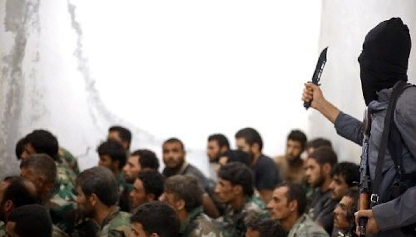 This undated image posted on Wednesday, Aug. 27, 2014 by the Raqqa Media Center of the Islamic State group, a Syrian opposition group, shows a fighter from the Islamic State group, armed with a knife and an automatic weapon, next to captured Syrian army soldiers and officers, following the battle for the Tabqa air base, in Raqqa, Syria.