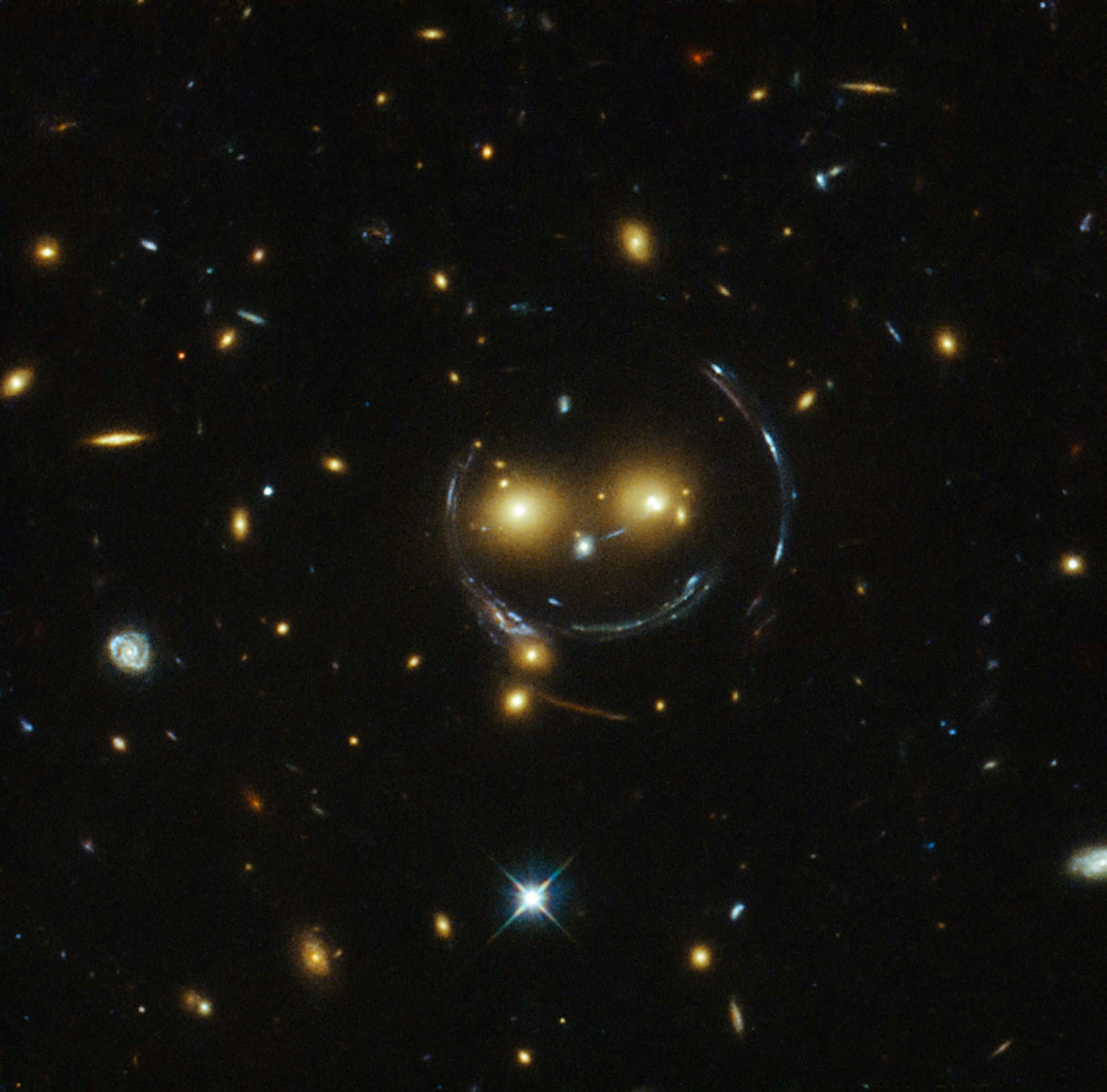 In the centre of this image, taken with the NASA/ESA Hubble Space Telescope, is the galaxy cluster SDSS J1038+4849 — and it seems to be smiling. You can make out its two orange eyes and white button nose. In the case of this "happy face", the two eyes are very bright galaxies and the misleading smile lines are actually arcs caused by an effect known as strong gravitational lensing. Galaxy clusters are the most massive structures in the Universe and exert such a powerful gravitational pull that t