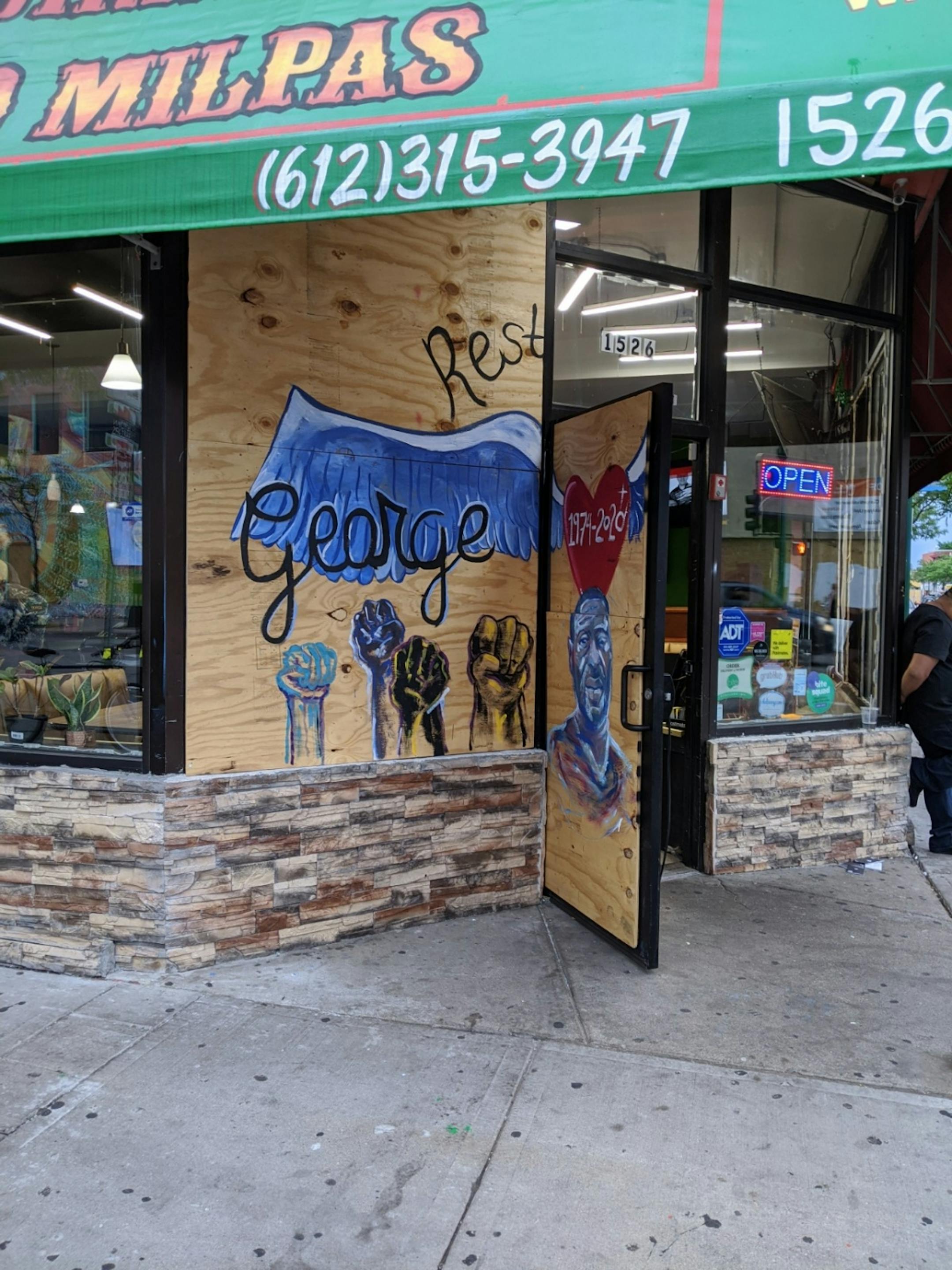 A mural over windows and door of Las Cuatro Milpas (1526 E Lake St., Minneapolis) portrays a blue-tinted George Floyd and fists raised in the air.