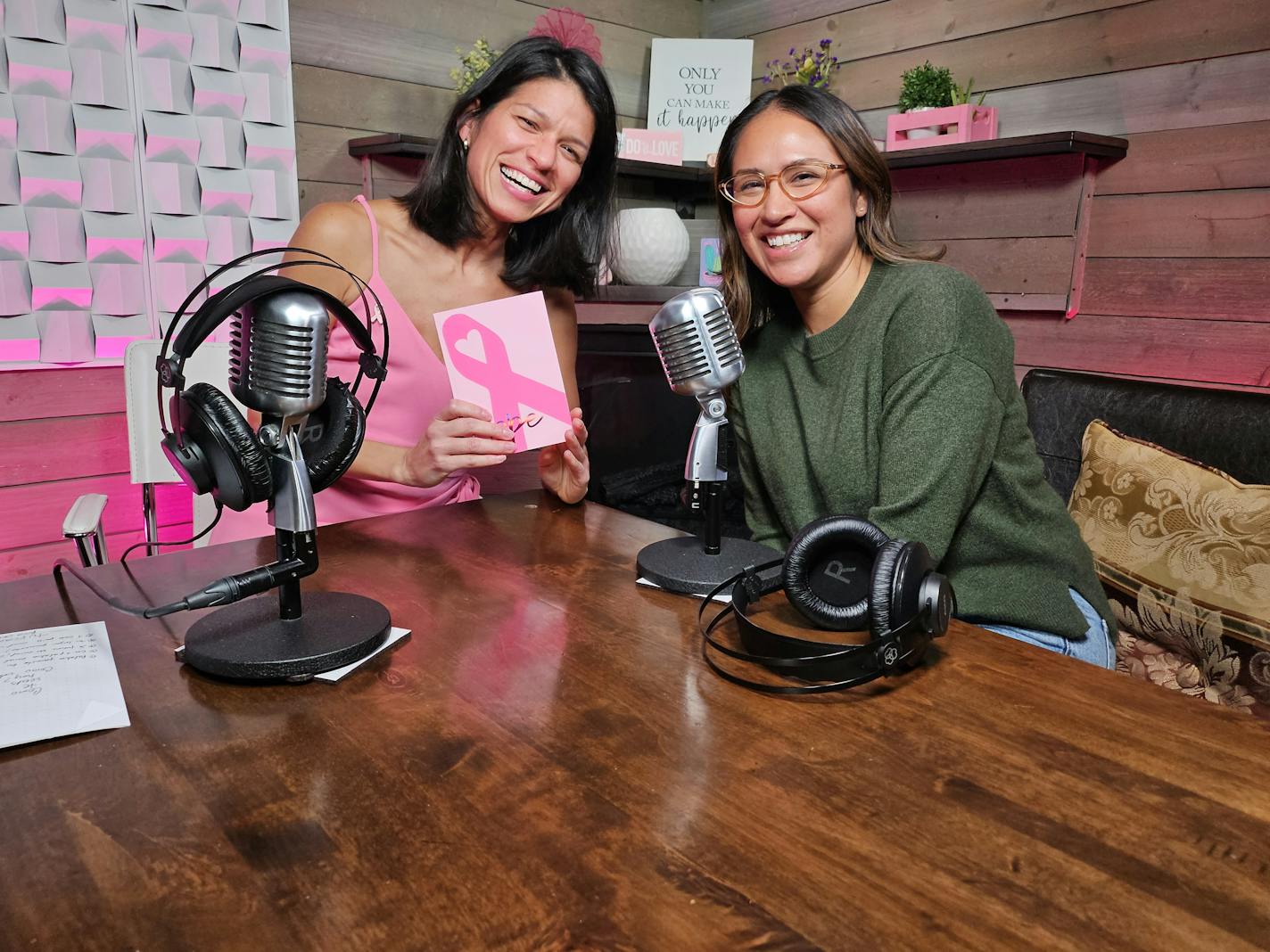 Two young women smile in a recording studio. The woman in a pink dress holds a sign with a pink ribbon, the sign for breast cancer awareness.