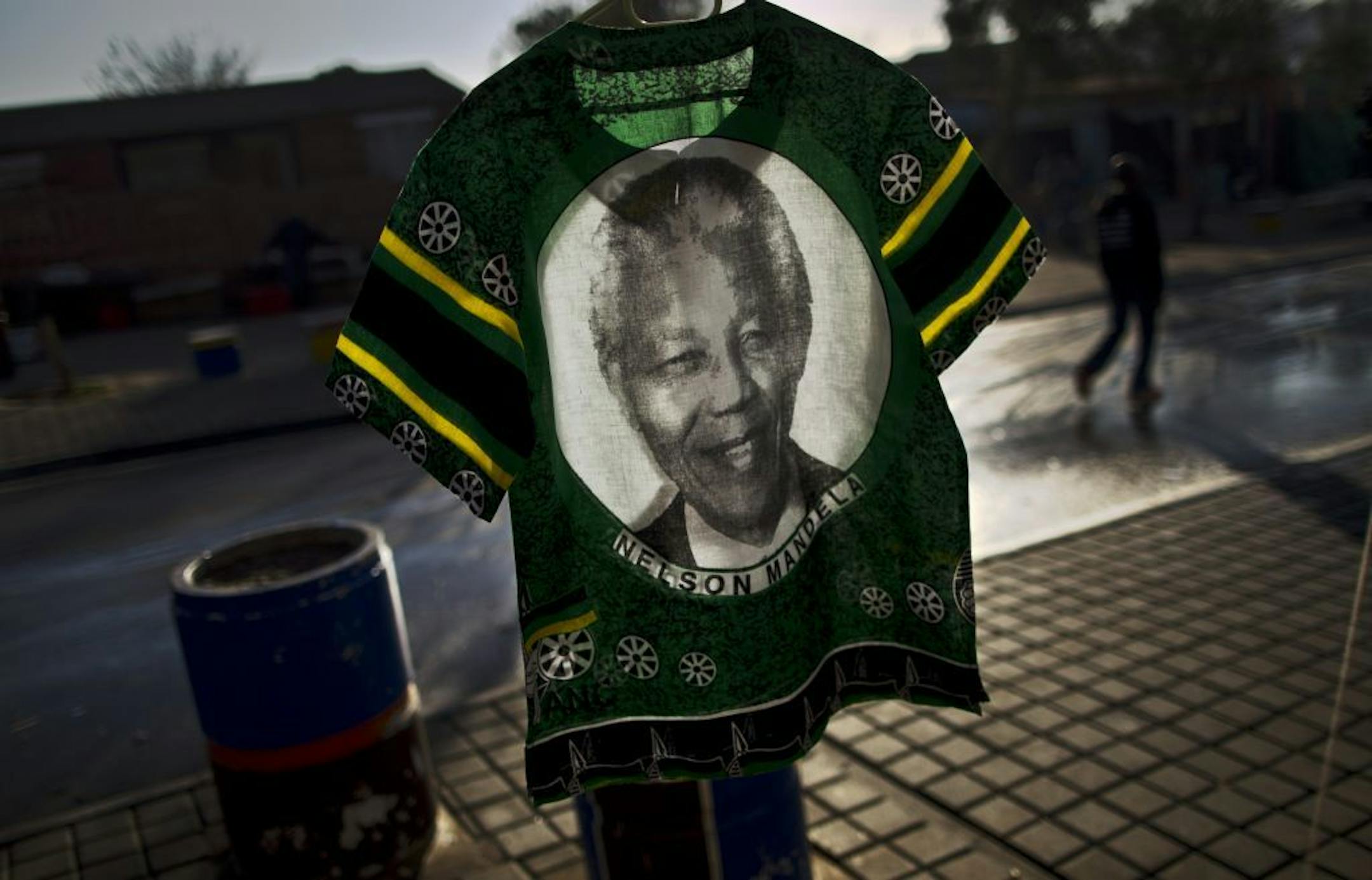 A South African man, right, walks past a T-shirt bearing the image of former South African President Nelson Mandela displayed for sale by a vendor, not pictured, in Soweto township on the outskirts of Johannesburg, South Africa, Monday, July 1, 2013. Former president Nelson Mandela remained in a critical condition at the Medi-Clinic Heart Hospital in Pretoria on Monday .