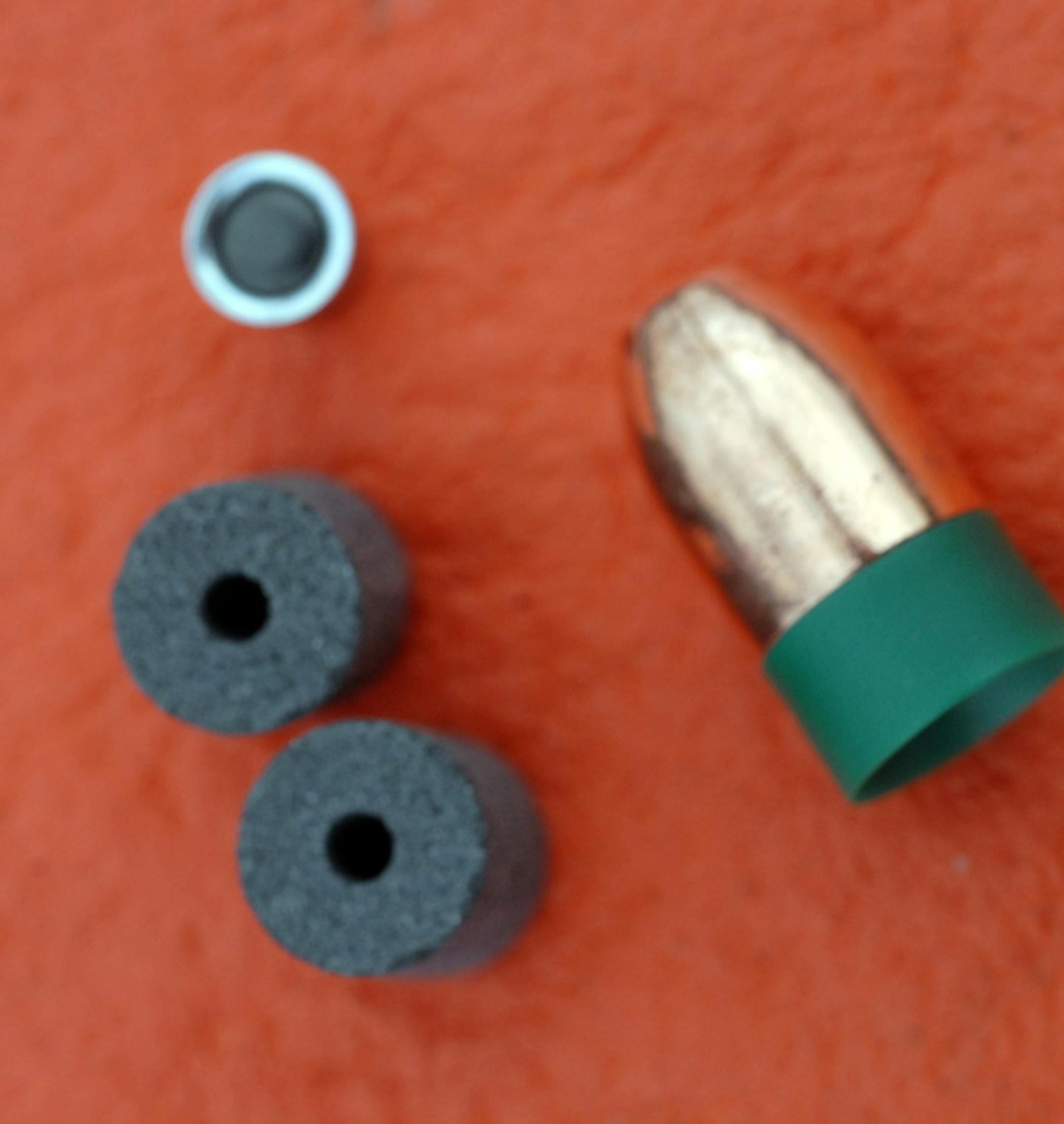 Many modern in-line muzzleloaders employ black powder substitutes like the two charges at left, foreground. A primer cap, top, provides ignition. A plastic-cased sabot-style bullet is at right.