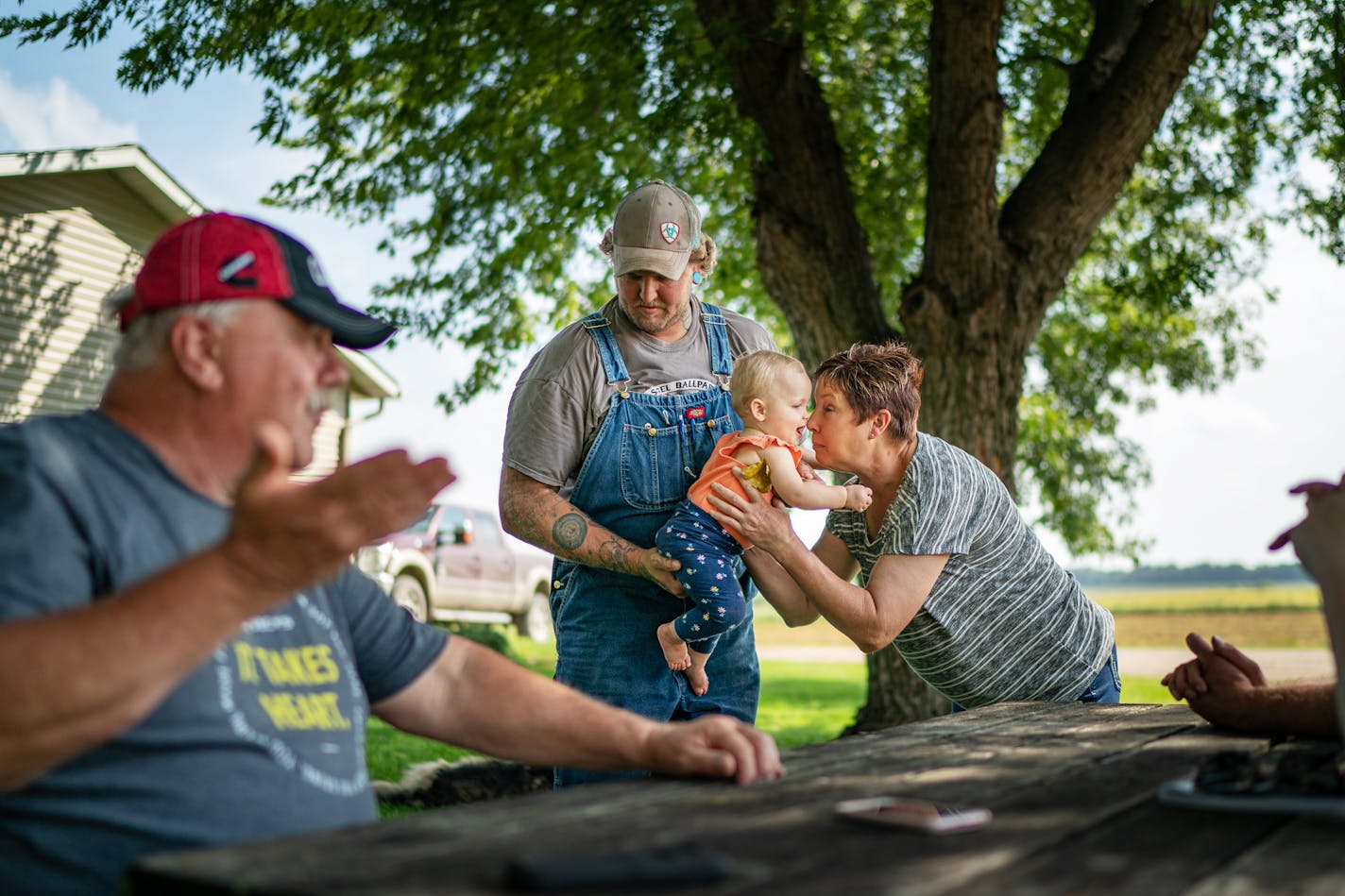 Mary Waibel cuddled granddaughter Elliott, 1, as son Jonathan Rewitzer and husband Tim, left, chatted on their farm in Courtland, Minn. Tim says trade had to be dealt with, but he doesn't like how Trump handled it.