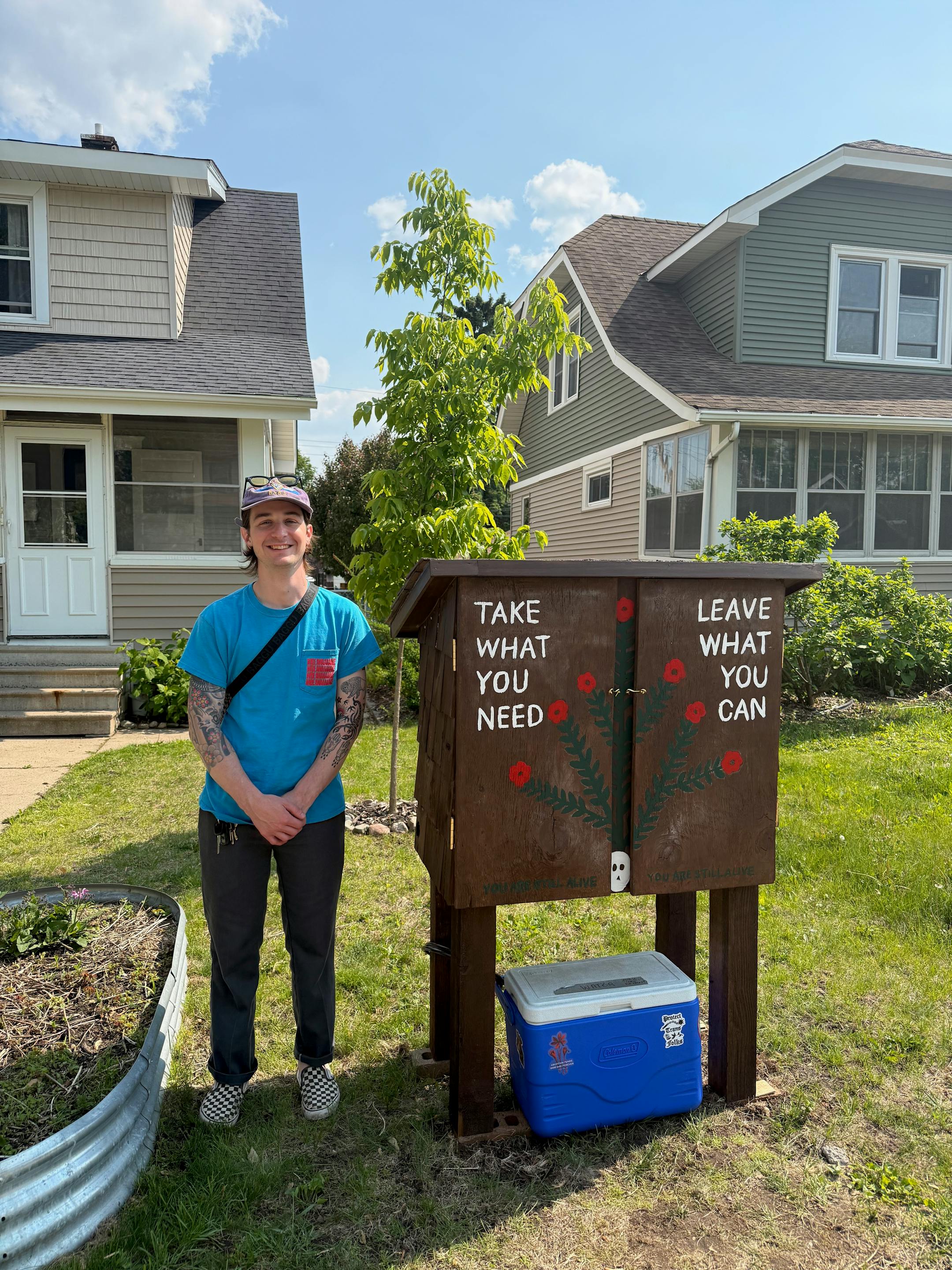 A man in his 30s stands next to a large wooden locker with a floral design painted on it, along with the words, "Take what you need. Leave what you can."