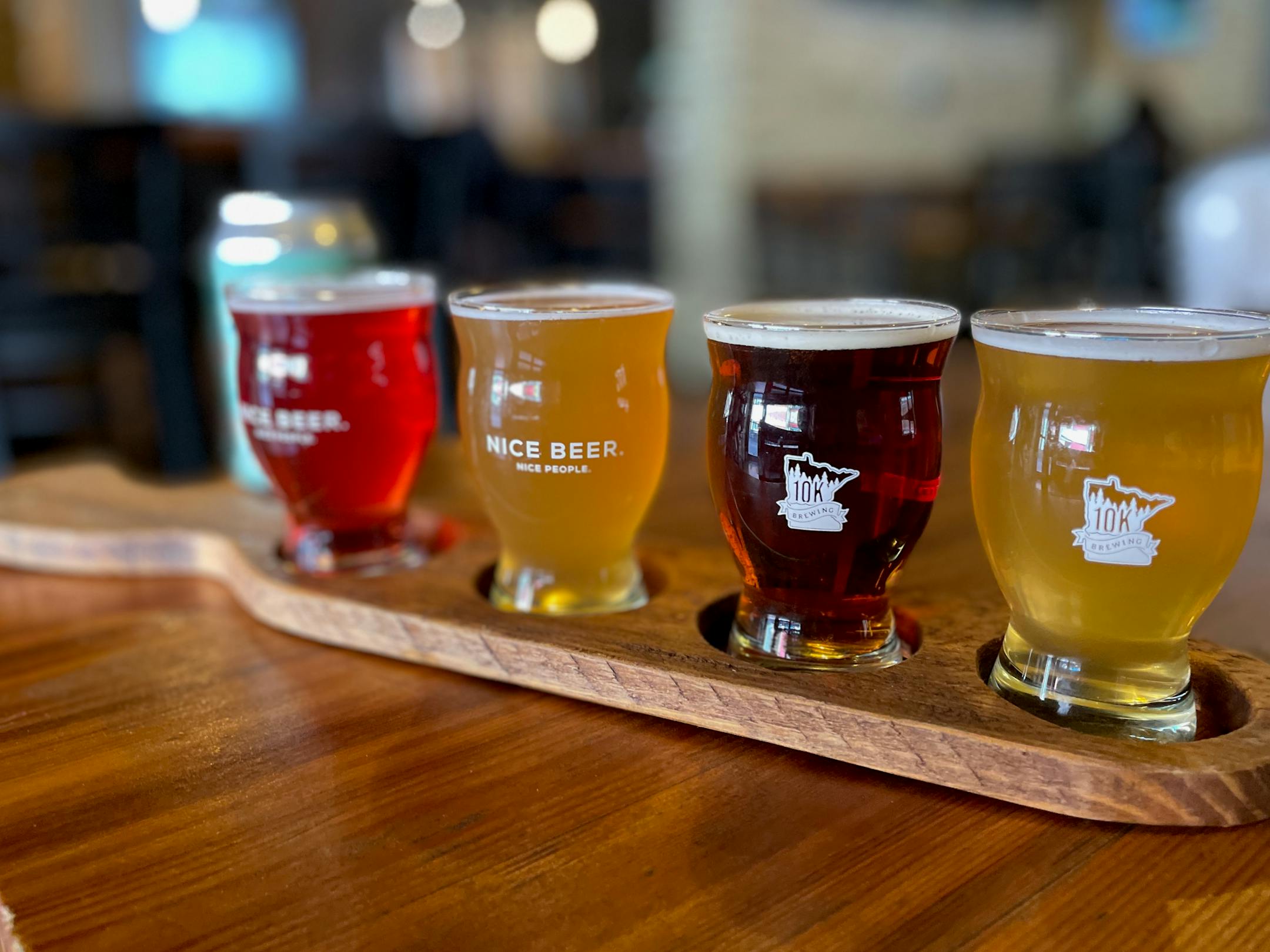 Four smaller glasses of varying colors of beer in a paddle shaped holder. The beer glasses sport the logo and slogan: Nice Beer. Nice People.