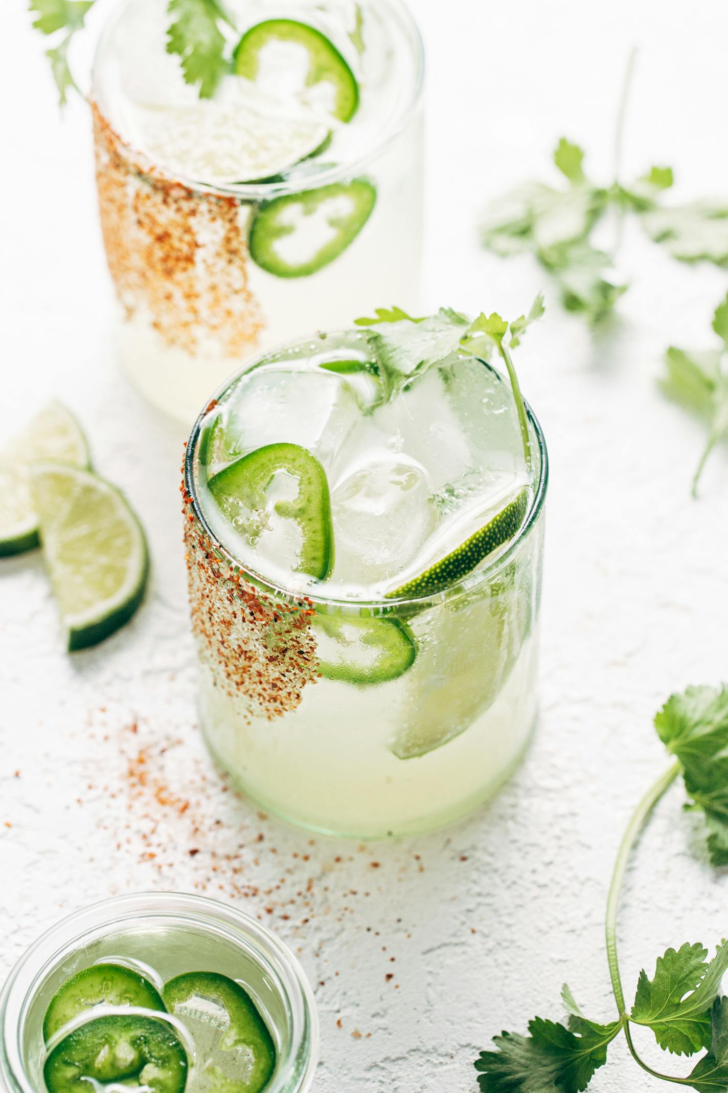 A cocktail with thin slices of jalapeno pepper and large ice cubs sits on a white countertop. It's dusted with red tajin spice.