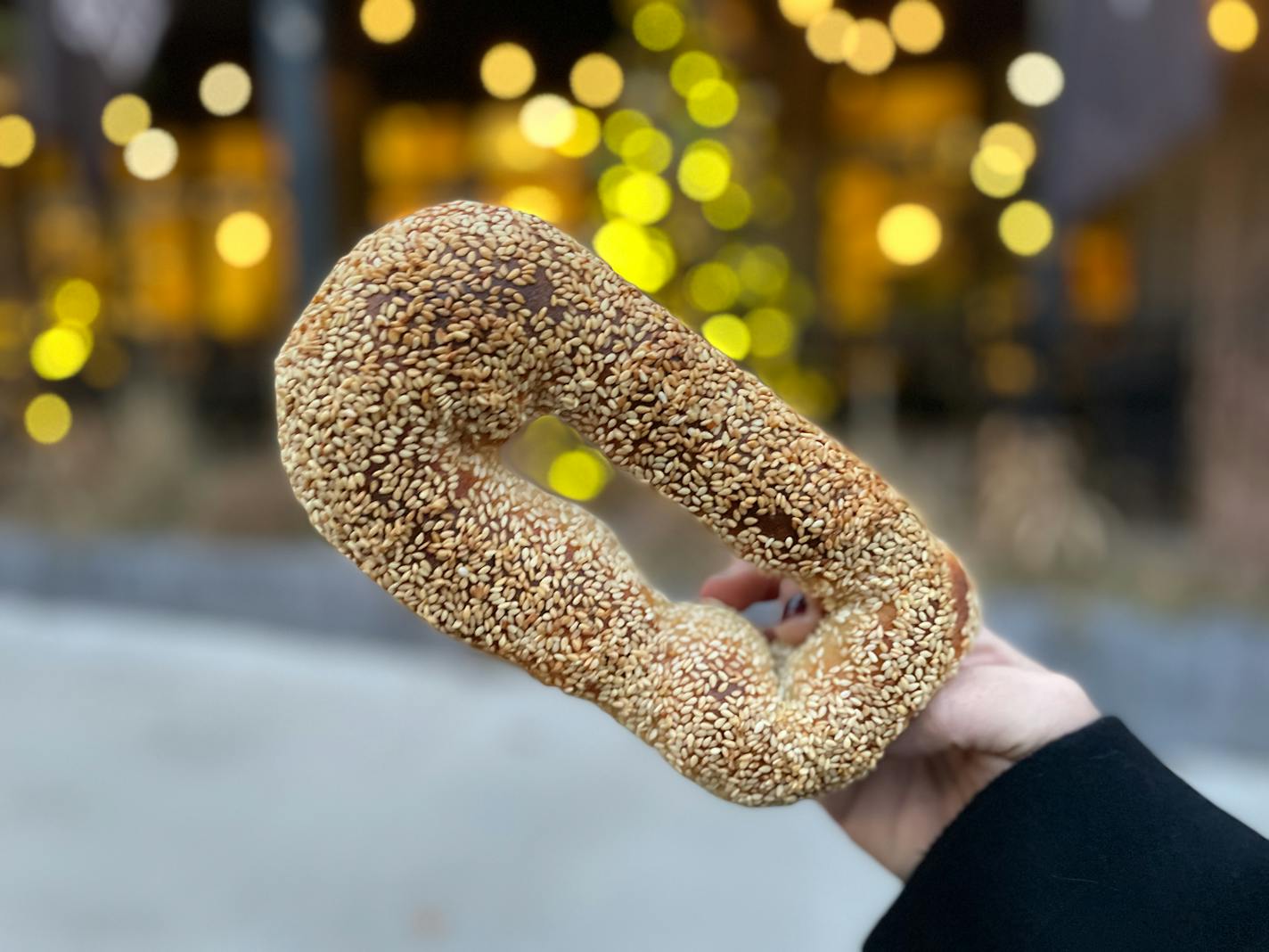 An oblong bagel studded with sesame seeds held in front of a twinkle light decorated building on Grand Avenue.