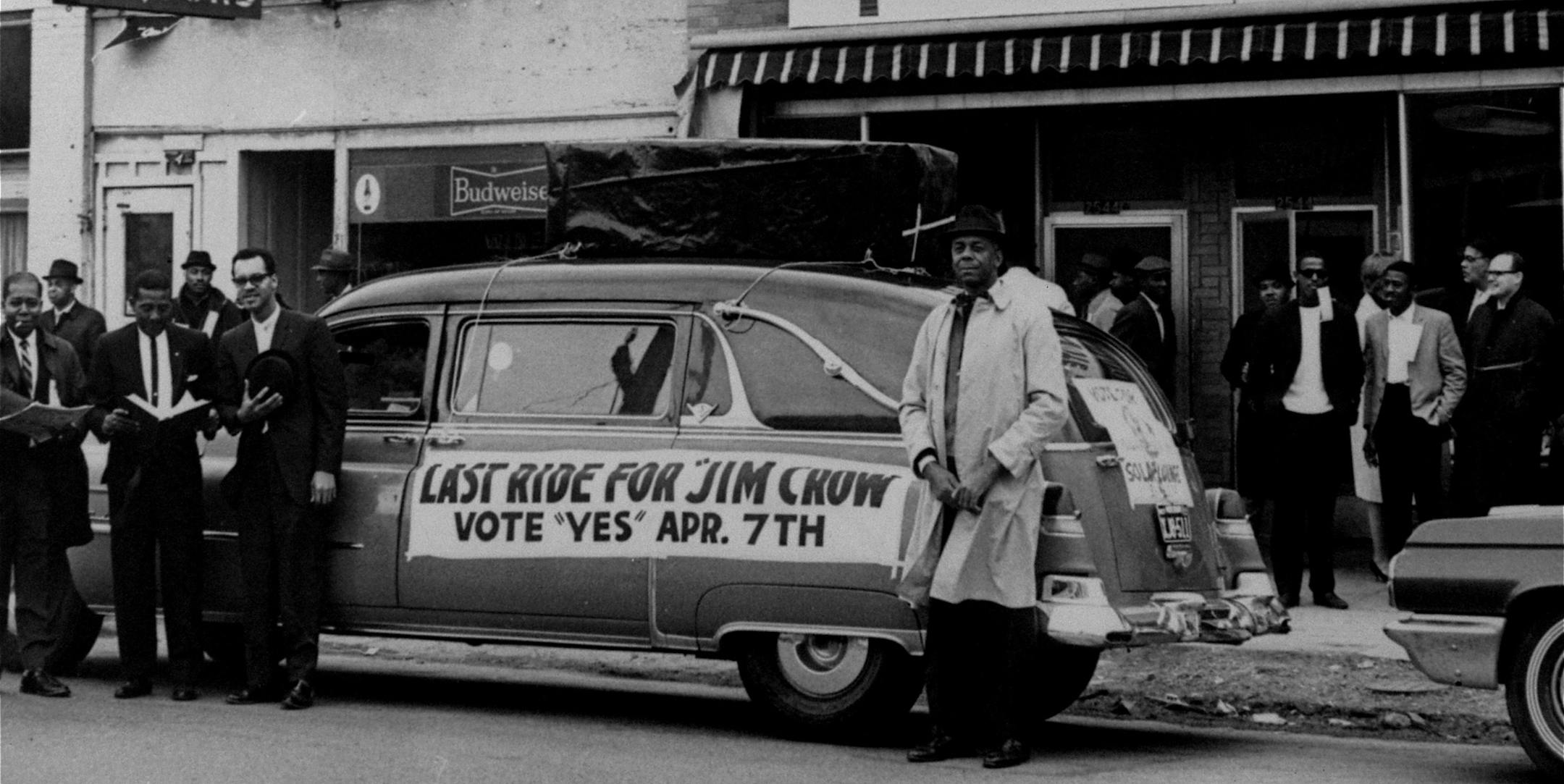 A hearse with a casket mounted on its top and a sign reading: Last Ride For Jim Crow, is parked in front of headquarters for workers favoring approval of Kansas City's Public Accommodations ordinance that was being voted on in a referendum election April 7, 1964. Three unidentified Negro campaign workers stand at left in front of hearse with heads uncovered and bowed in mock sorrow for Jim Crow. A heavy turnout was reported in the vote for the ordinance that makes it illegal for owners of virtua