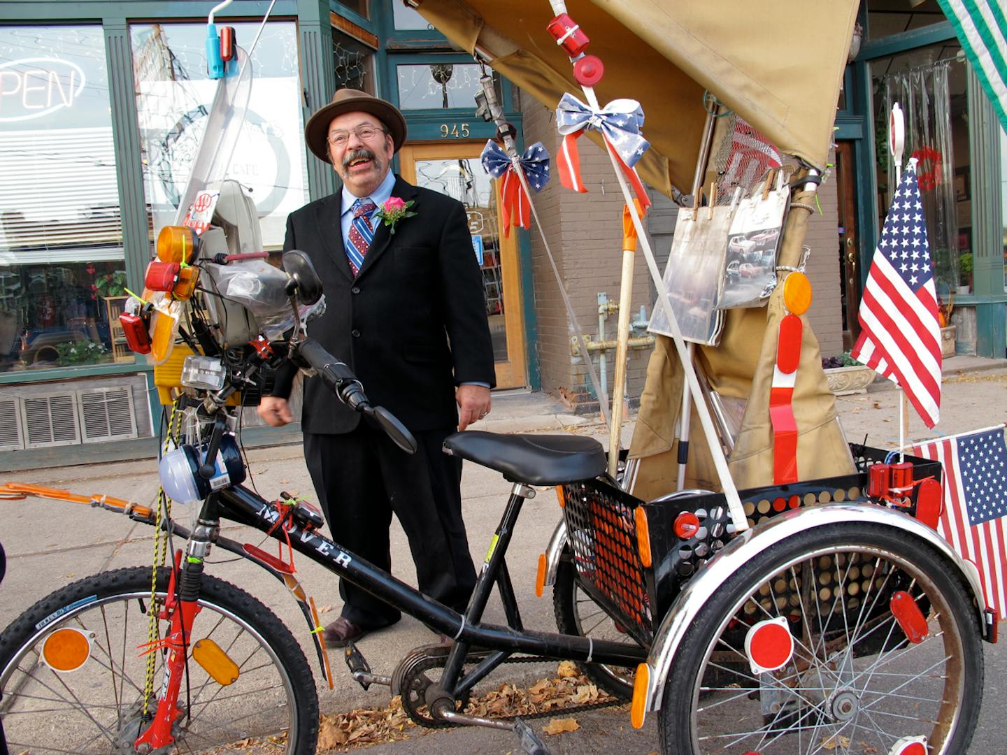 Carl Bentson wears a suit and pink boutonniere as he stands and smiles behind his tricycle outside a storefront.