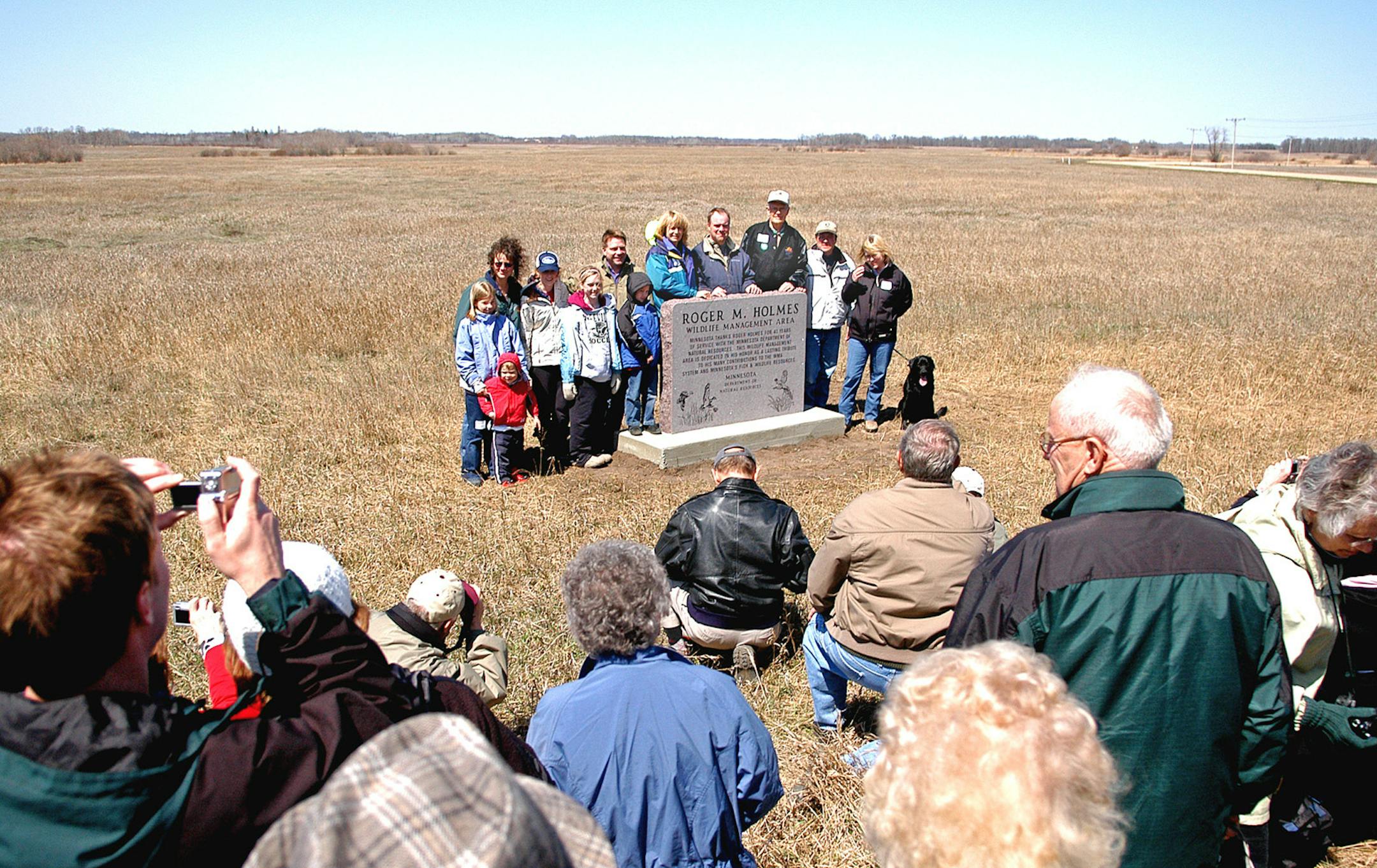 Retired Department of Natural Resources fish and wildlife division director Roger Holmes, behind monument in dark jacket and white cap, was honored in Douglas County, near Alexandria, Minn., on Saturday.