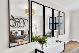 A row of five vertical mirrors with black frames on a white wall reflects a living room.