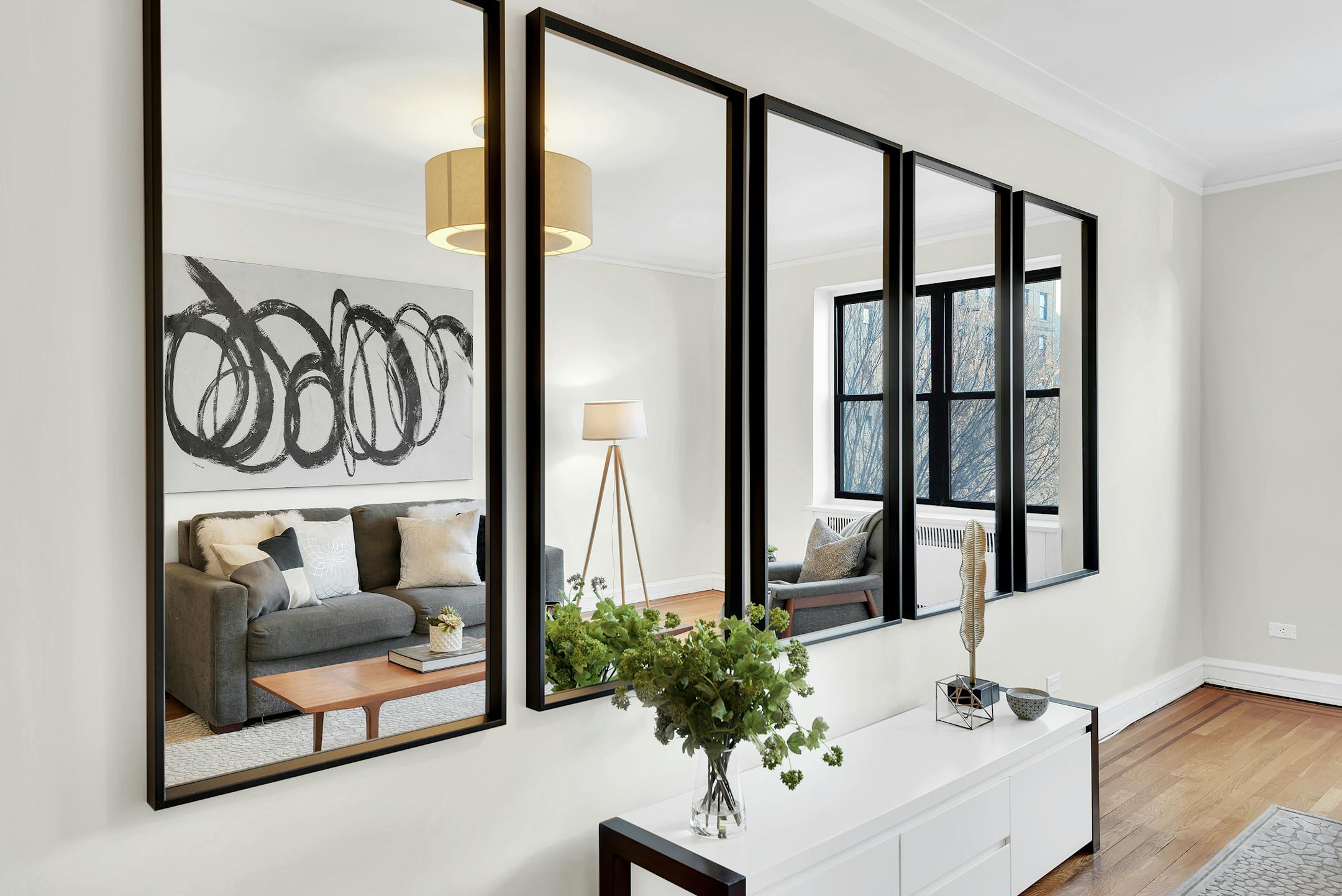 A row of five vertical mirrors with black frames on a white wall reflects a living room.