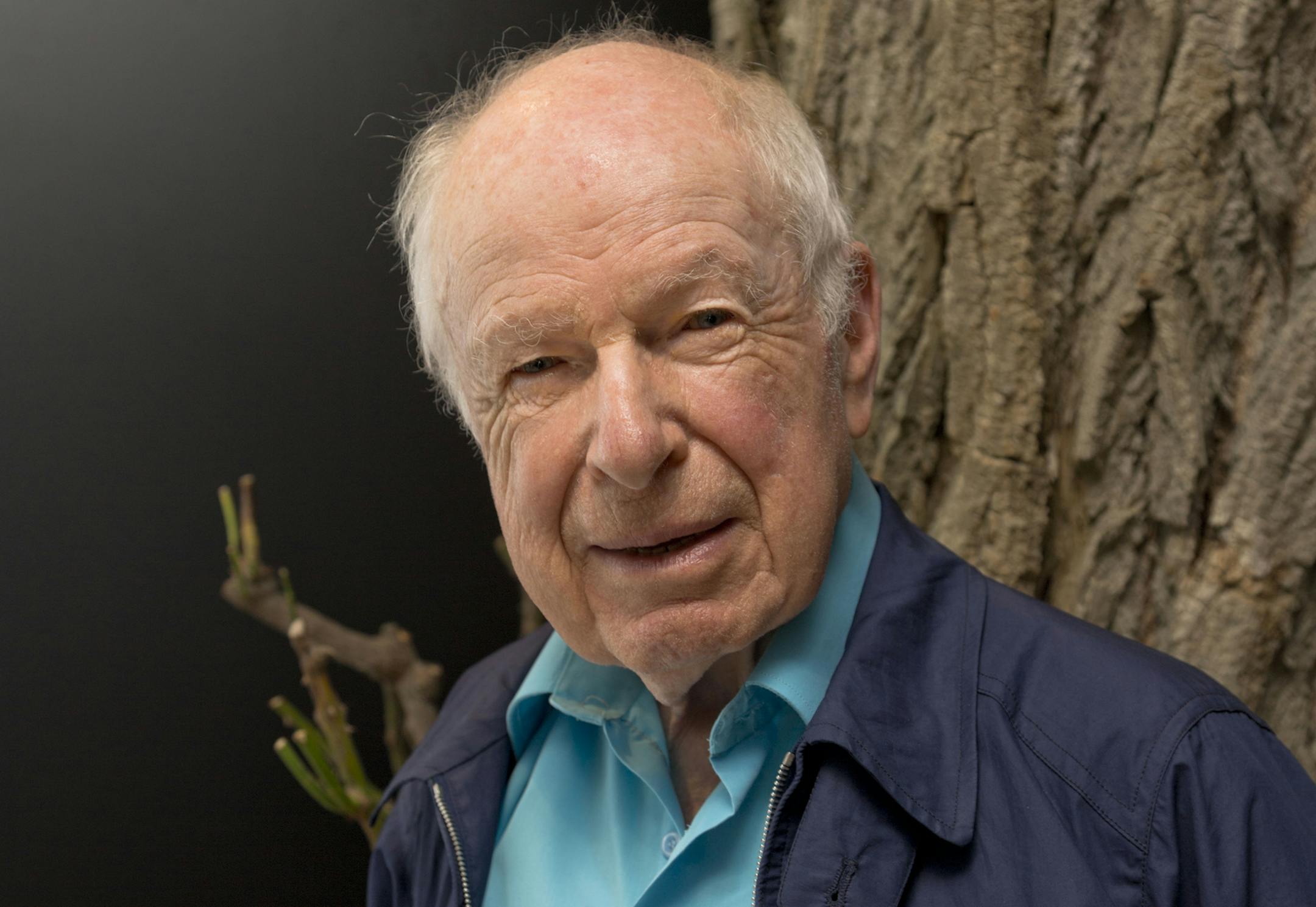 Closeup of Peter Brook wearing a turquoise polo shirt and blue collared jacket and standing in front of tree