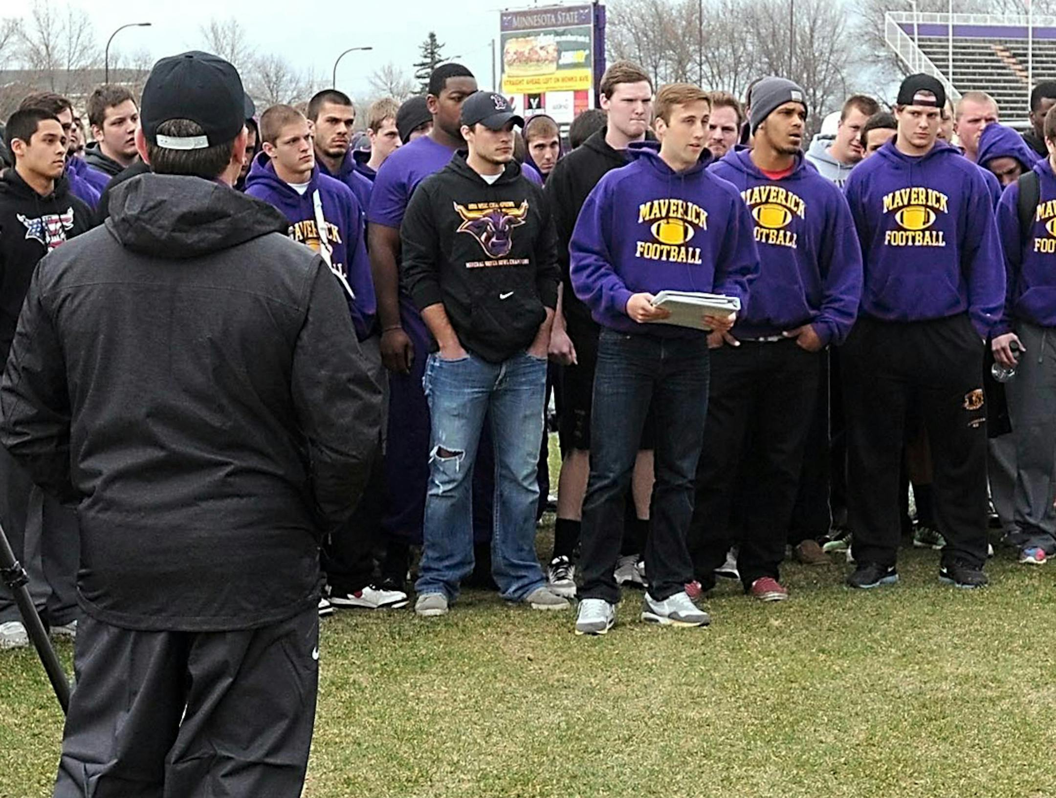 Reinstated Minnesota State, Mankato football coach Todd Hoffner, foreground, listens to a statement read by football player Samuel Johnson as the rest of the team stands by Wednesday, April 16, 2014 in Mankato, Minn. An arbitrator ruled last week that Hoffner was wrongfully dismissed in the wake of a child porn charges that were ultimately dropped. Players came out for spring practice Wednesday but weren't in uniform. In the statement, players said they want interim coach Aaron Keen back. (AP Ph