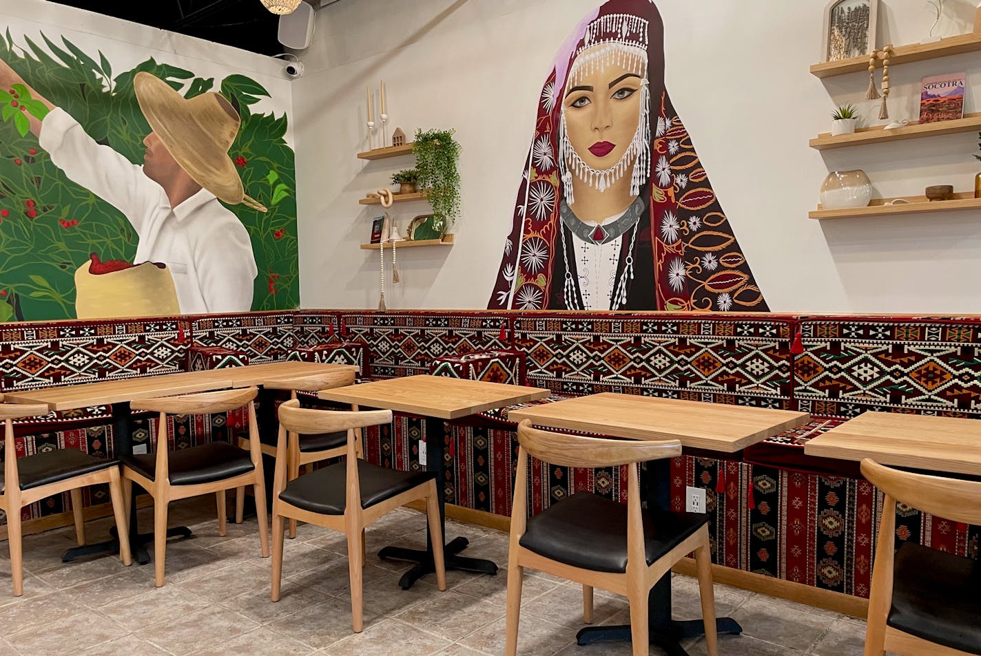 The image shows the pattered cloth seats along the wall and a few tables below murals of a woman in traditional dress and a man harvesting coffee berries.