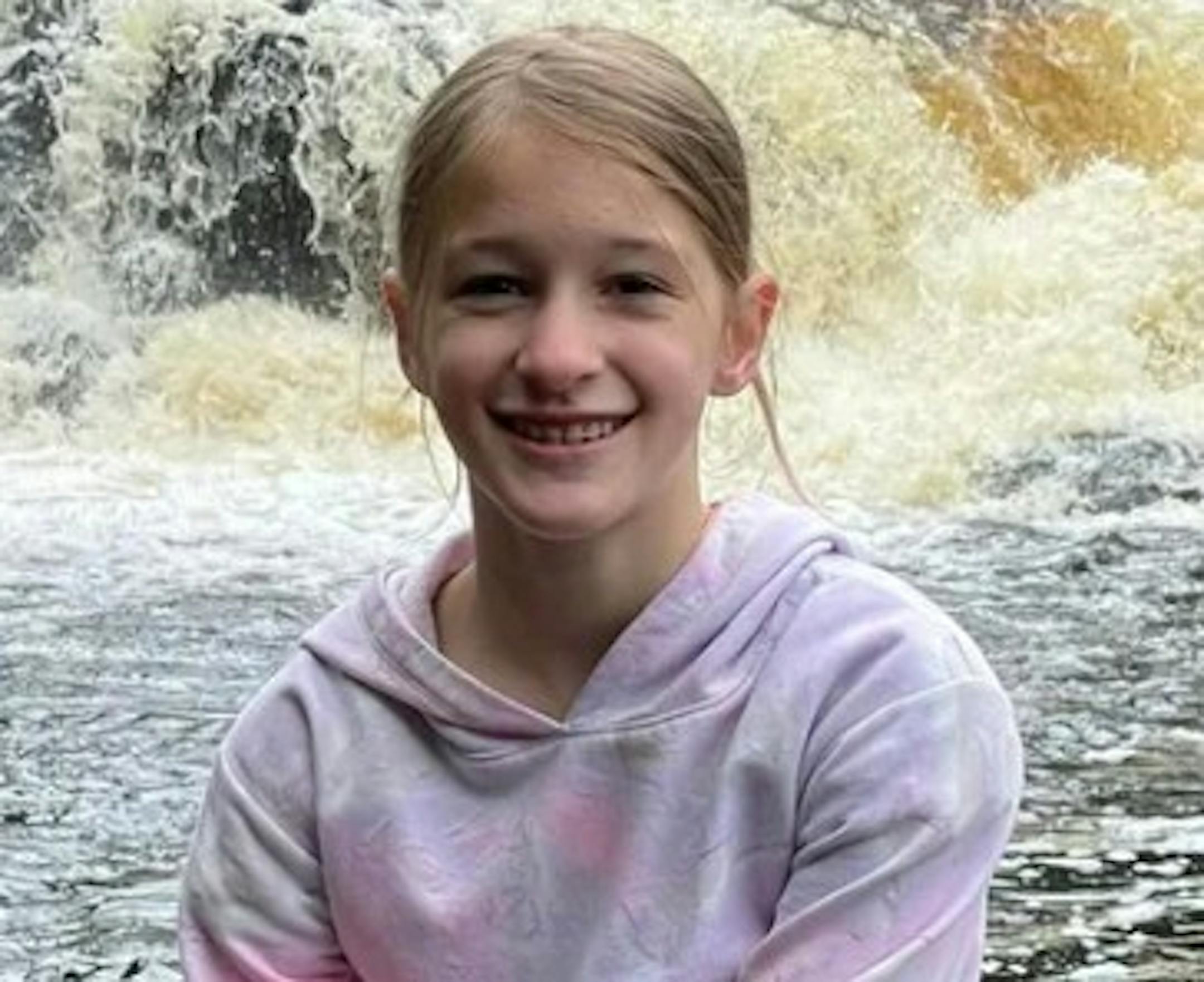 A blond girl in a hoodie smiles in front of what looks to be the base of a waterfall.