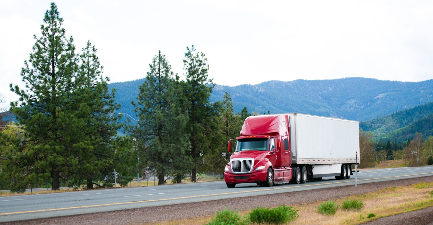 Red big rig modern shiny semi-truck with white dry van trailer with aerodynamic skirt move on straight divided interstate highway I-5 in California with green trees and mountains on background