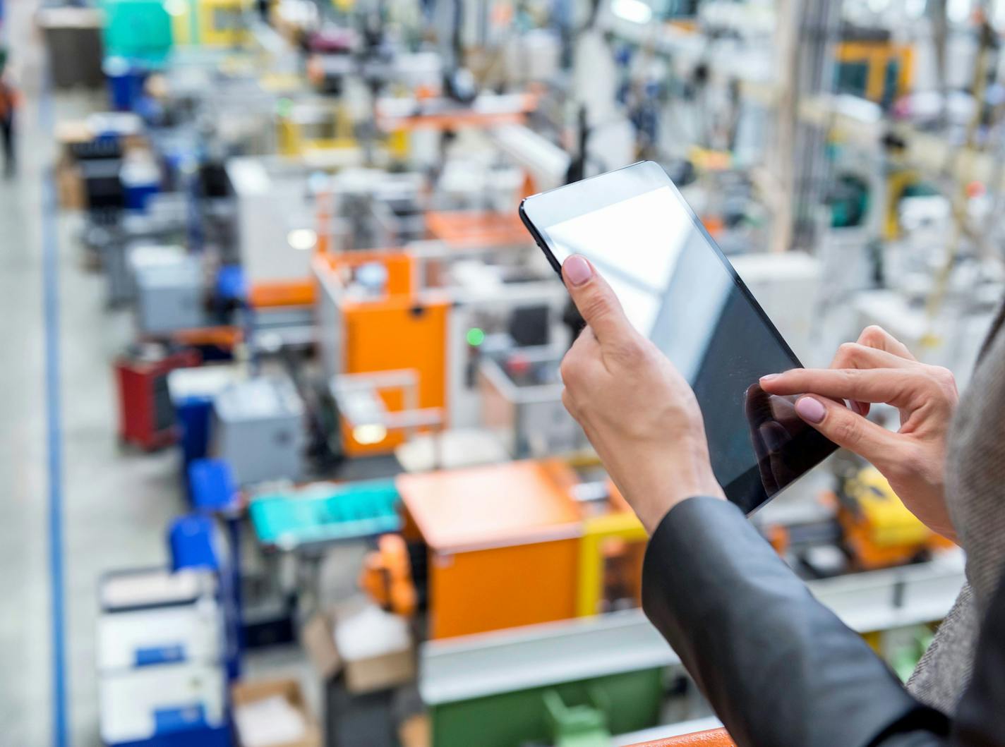 Horizontal color image of businesswoman - unrecognizable person - working with digital tablet in large futuristic factory. Woman standing on top of a balcony, holding touchpad and checking inventory of a manufacturing company on touchscreen tablet. Focus on businesswoman's hands holding black tablet, futuristic machines in background.