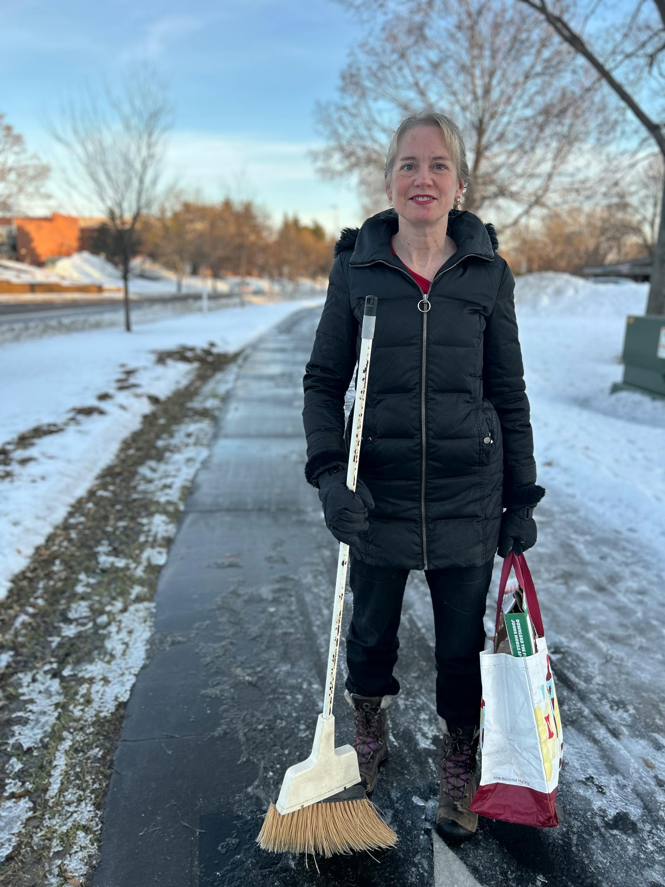A woman in a parka stands along a walking path with a broom and a tote bag of soiled cardboard and other organic recycling.