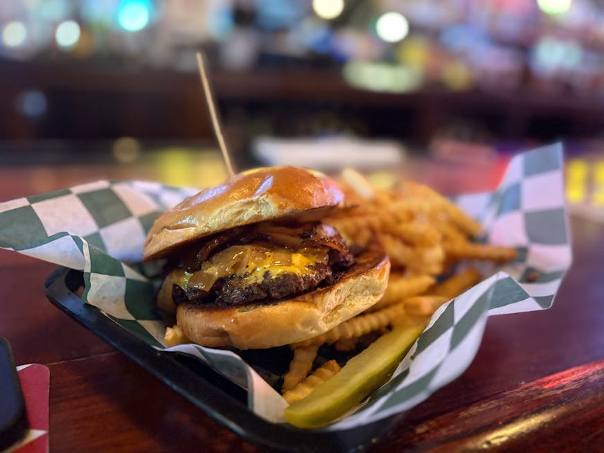 In a basket on an old bar is a burger with cheese, carmelized onions on a bun with a toothpick through it and fries on the side.