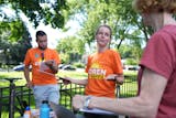 Woman in orange shirt with clipboard talks during a meeting of political volunteers in a leafy park.