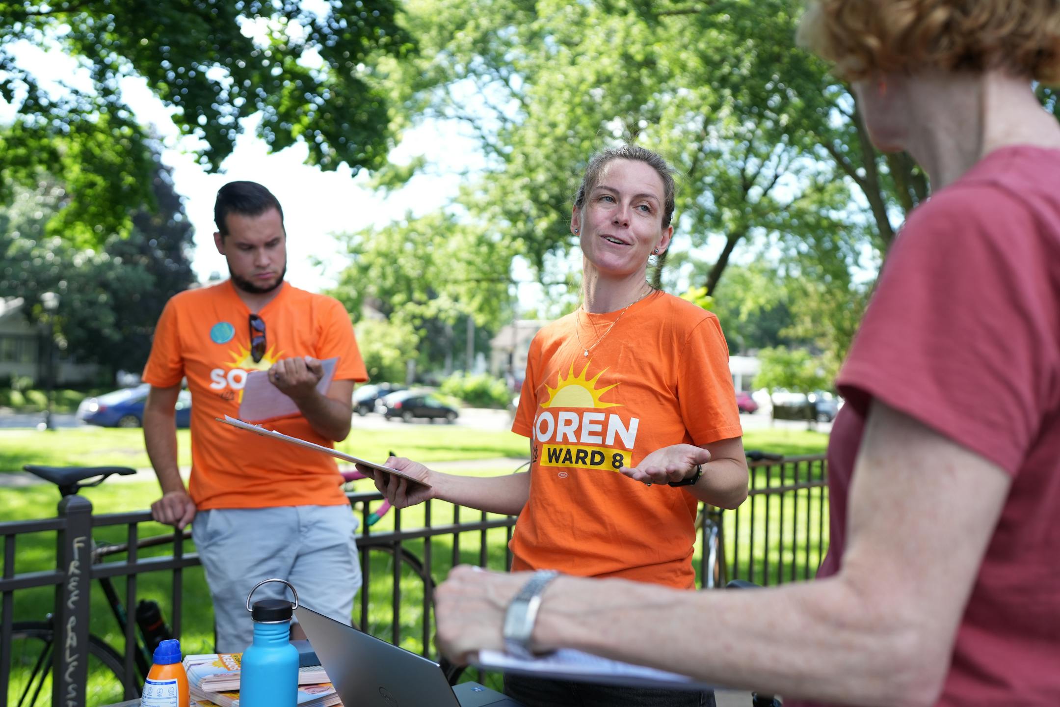 Woman in orange shirt with clipboard talks during a meeting of political volunteers in a leafy park.