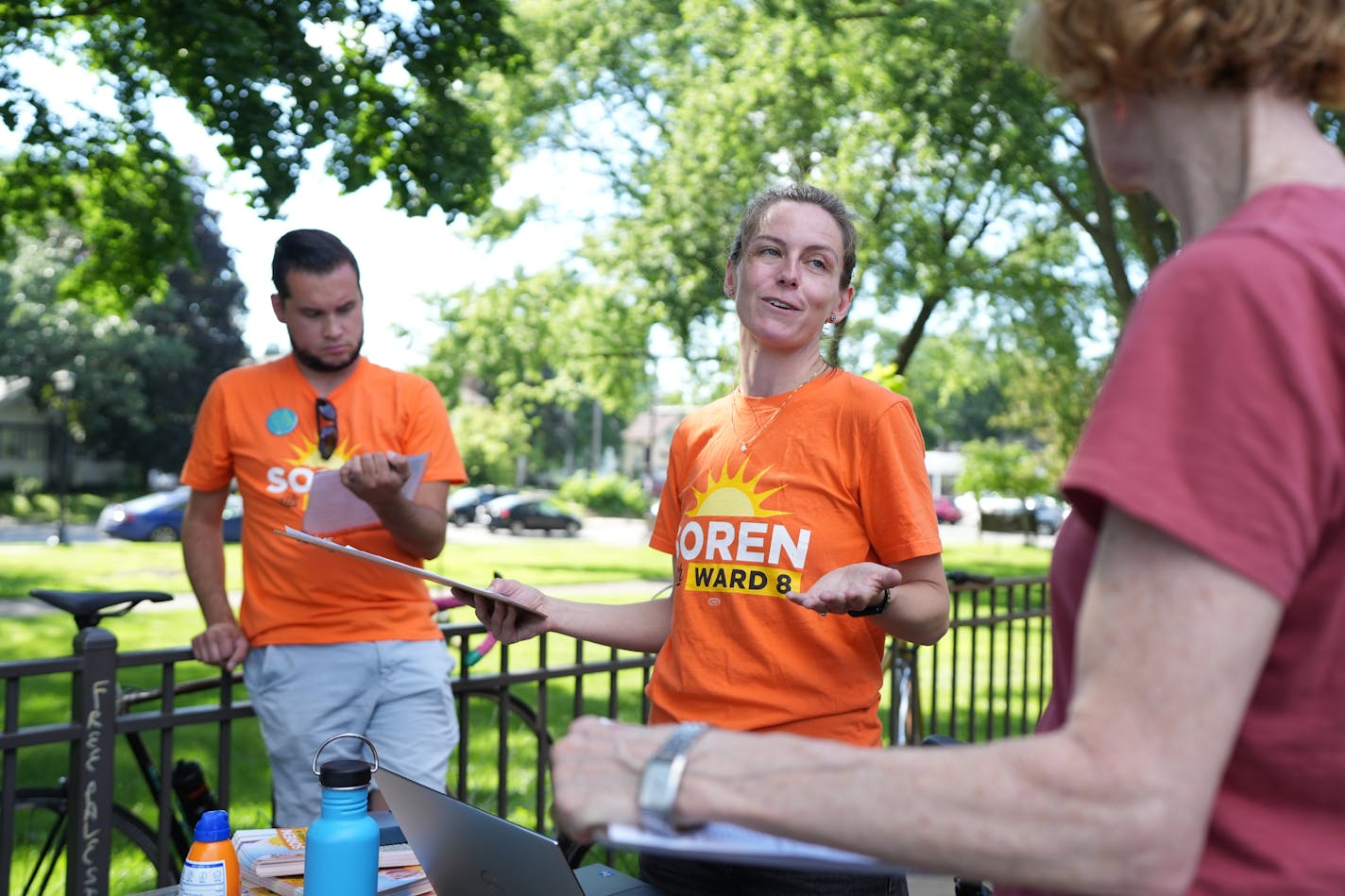 Woman in orange shirt with clipboard talks during a meeting of political volunteers in a leafy park.