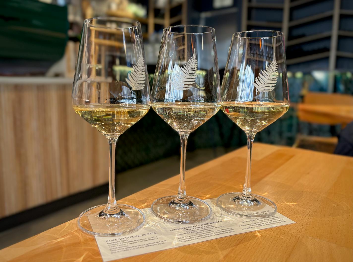 A white wine flight on a light wood table with a deep green/mermaid tail tiled bar in the background.