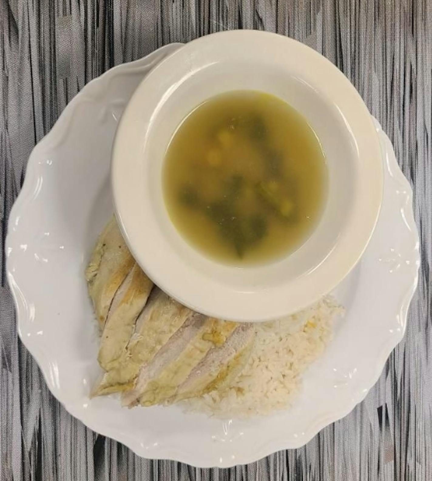 A bowl of yellow broth and green herbs sits on a plate that also includes white rice and sliced cooked chicken.