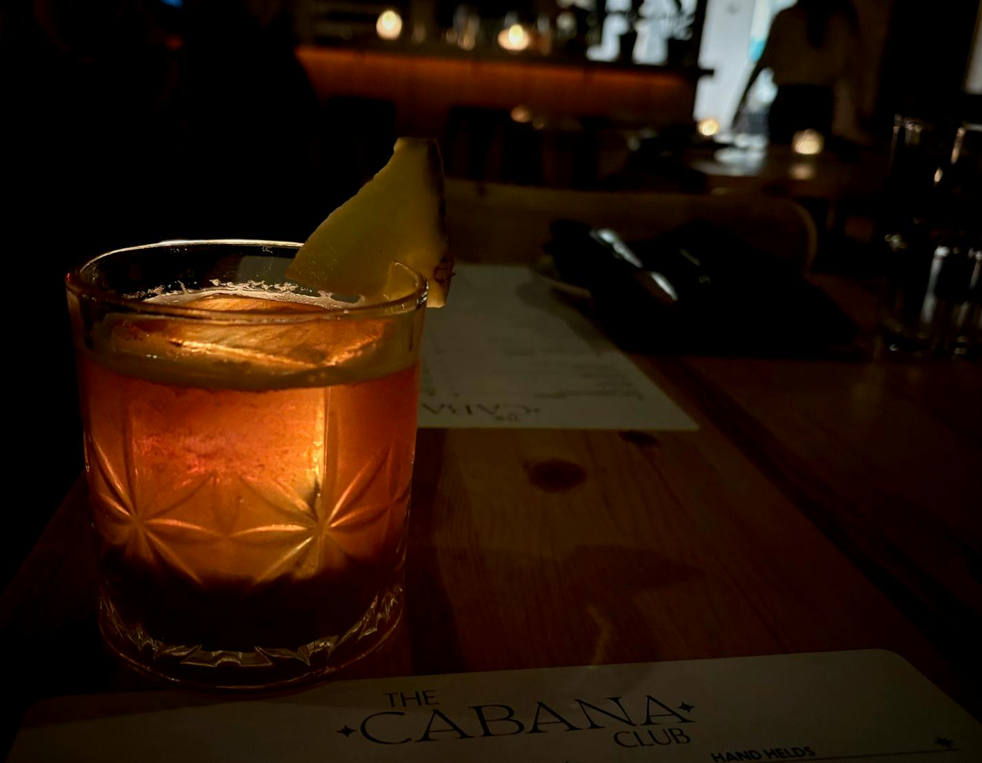 A lowball cocktail with a large, square ice cube is back lit by a candle, illuminating the amber liquid inside. Beneath it is a paper menu that says, "The Cabana Club."