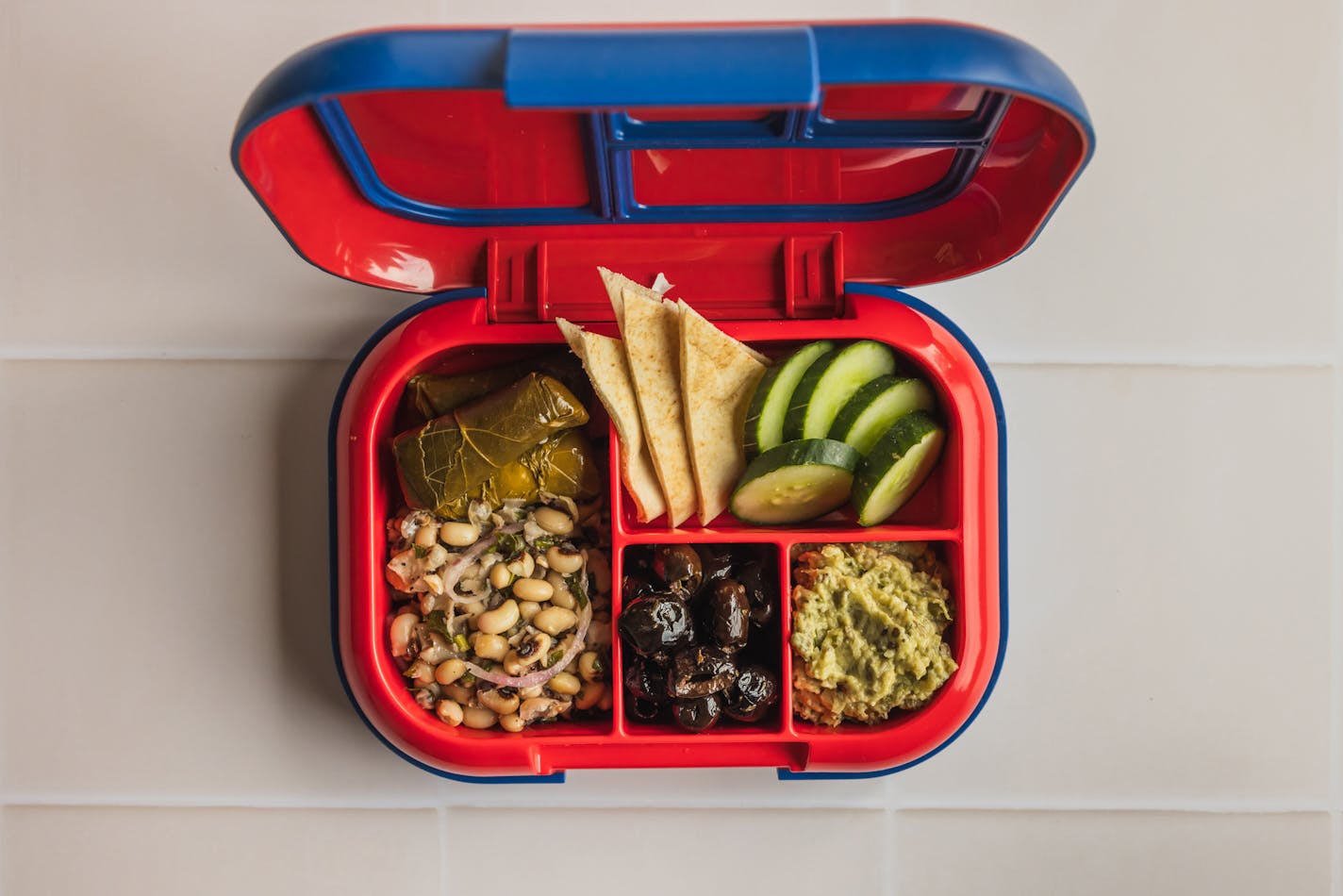 A bright red and blue bento box lunch container with Dolmas and bean salad, pitas and cucumbers, eggplant dip and marinated olives.