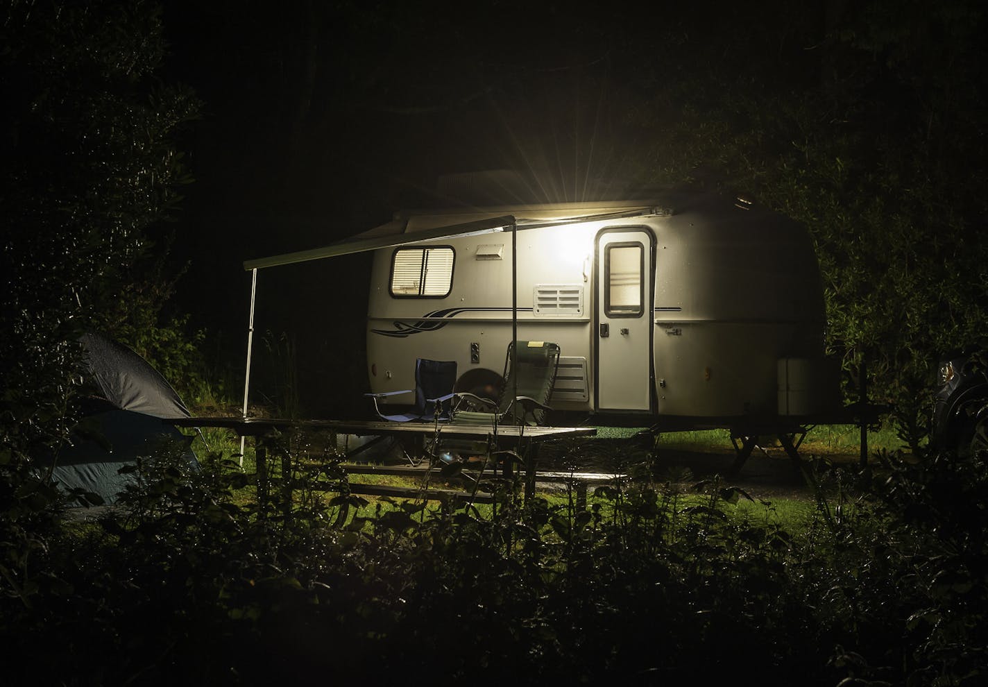 Compact travel trailer glowing with warm light through windows in a forest RV park in the Pacific Northwest of the USA.