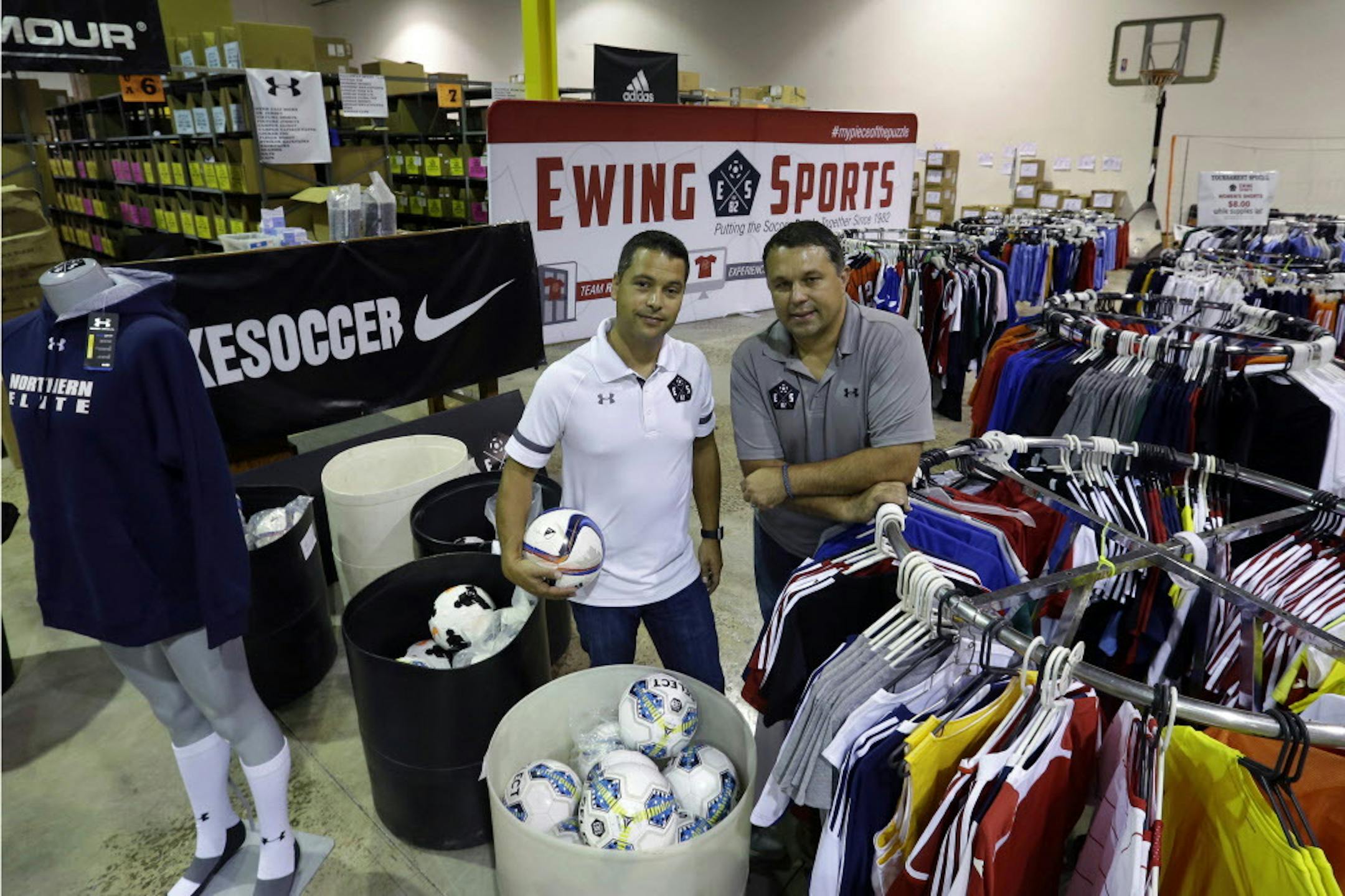 In this July 29, 2015 photo, Chris Goodrich, left, and Les Korsos, pose with sale racks stocked with soccer uniforms and gear at a warehouse of their company, Ewing Sports, that sells mostly soccer uniforms to school and other teams, in Ewing, N.J., Key employees at the company, Goodrich and Korsos bought Ewing Sports from their retiring bosses in July. They are part of a trend that is picking up momentum as sales of businesses increase. (AP Photo/Mel Evans)