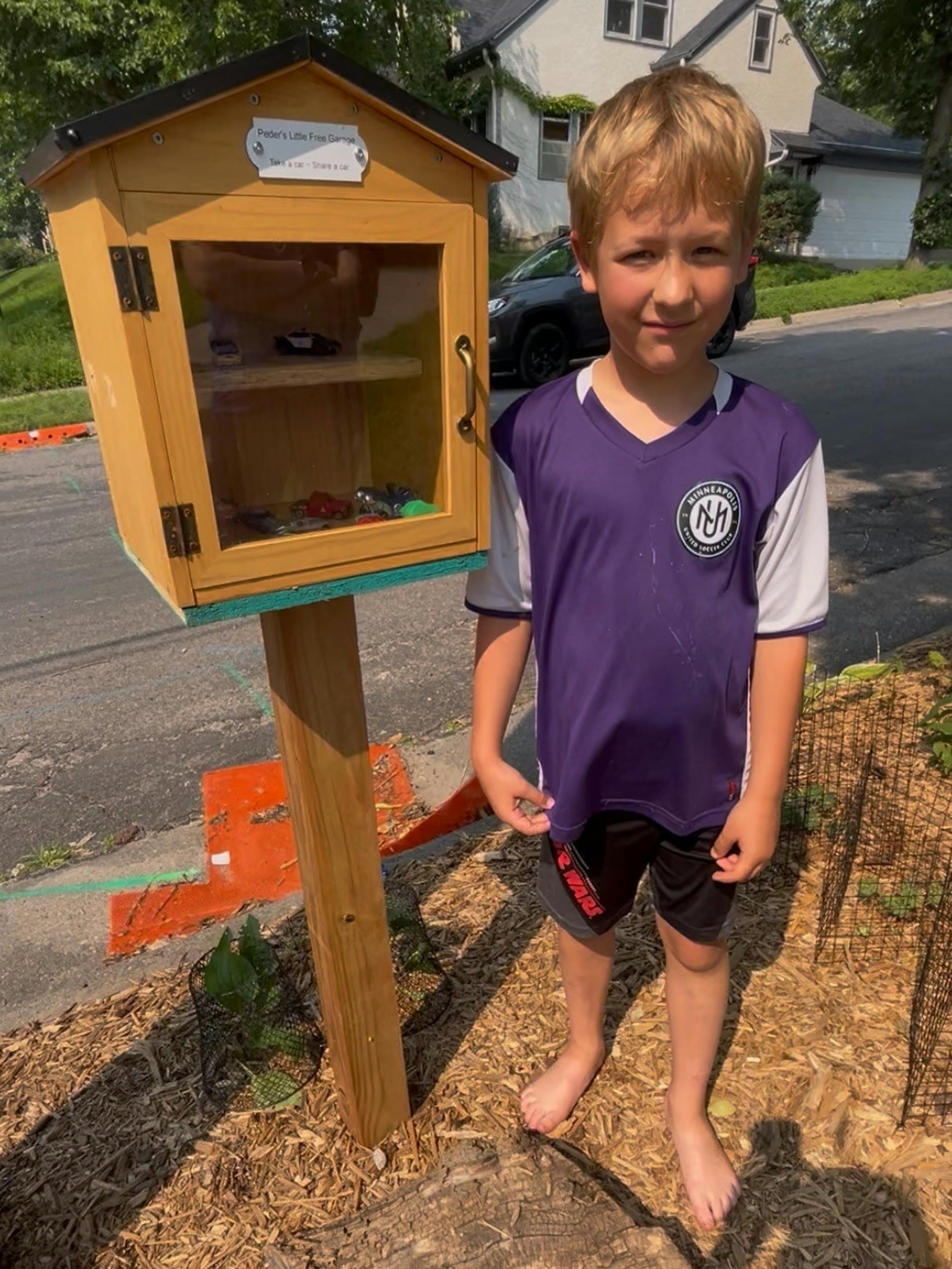 In a boulevard of a residential neighborhood, a barefooted 8-year-old boy wearing a soccer jersey stands by a little free library containing matchbox cars.