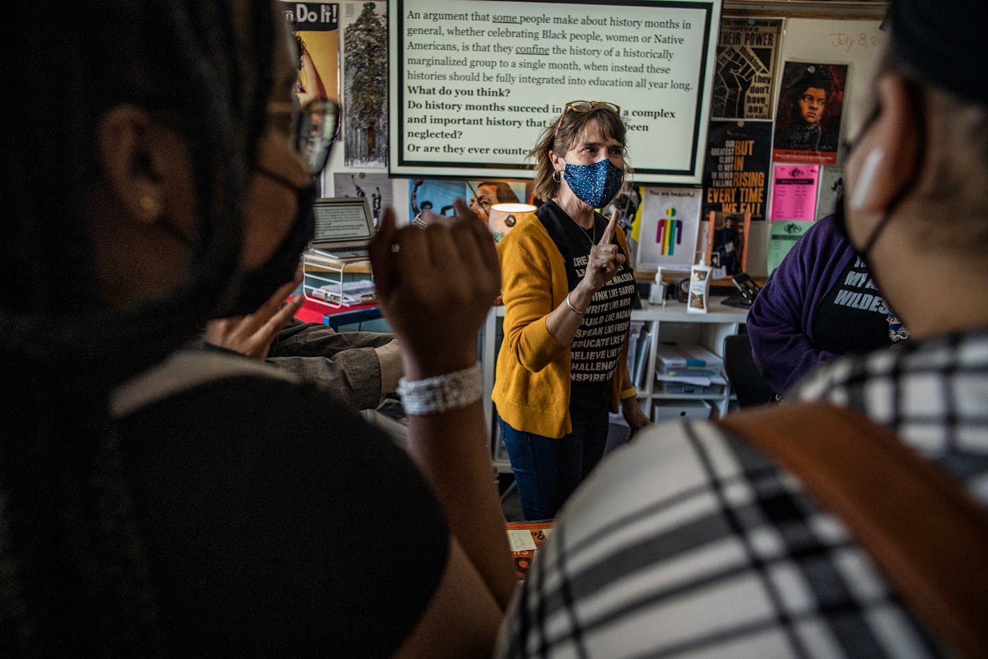 Two students stand in the foreground with their backs to the camera as a masked woman facing them points up