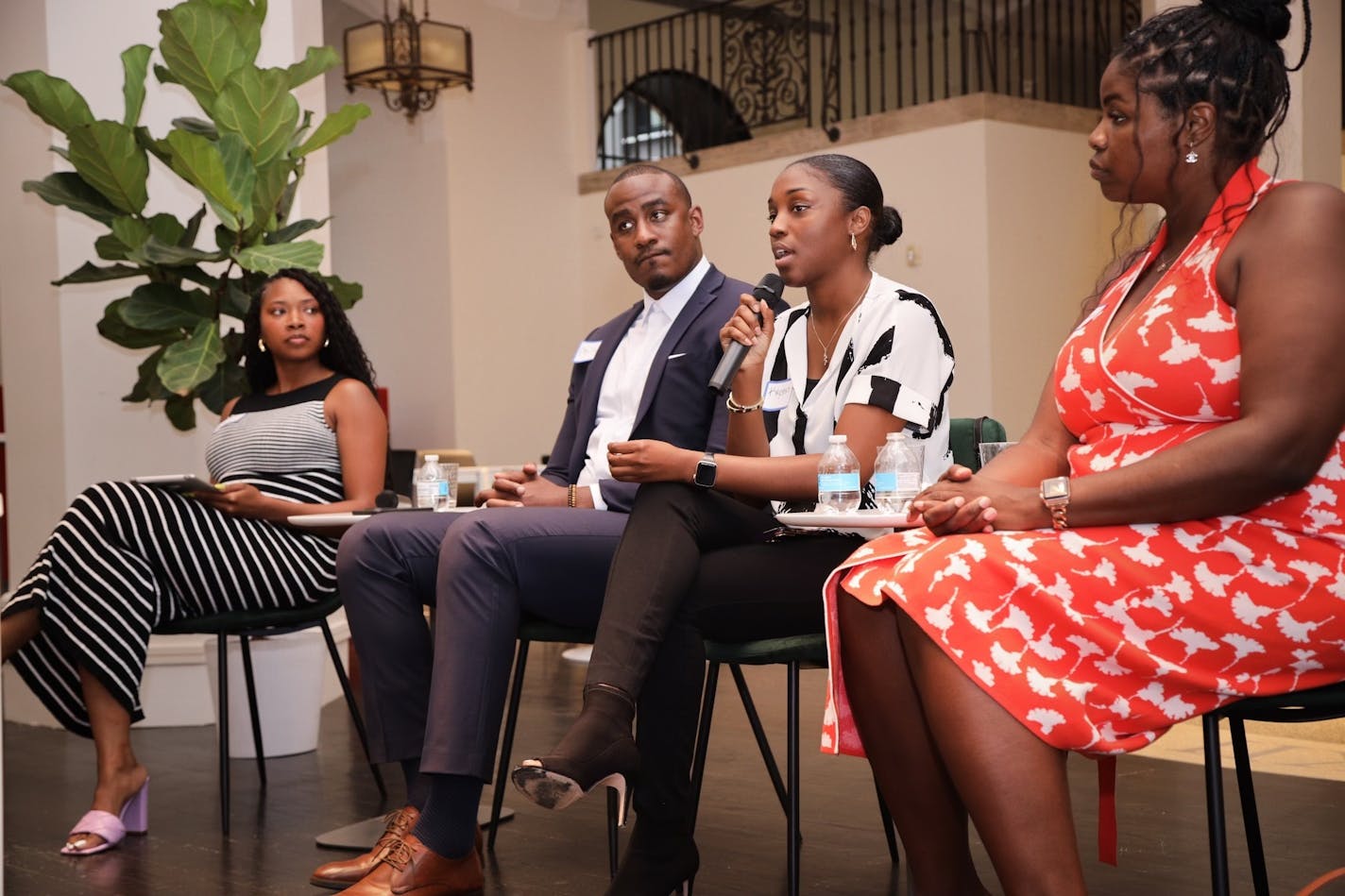 Four young Black professionals in suits and dresses sit on chairs. One woman holds a microphone and is speaking to the audience while the others listen.