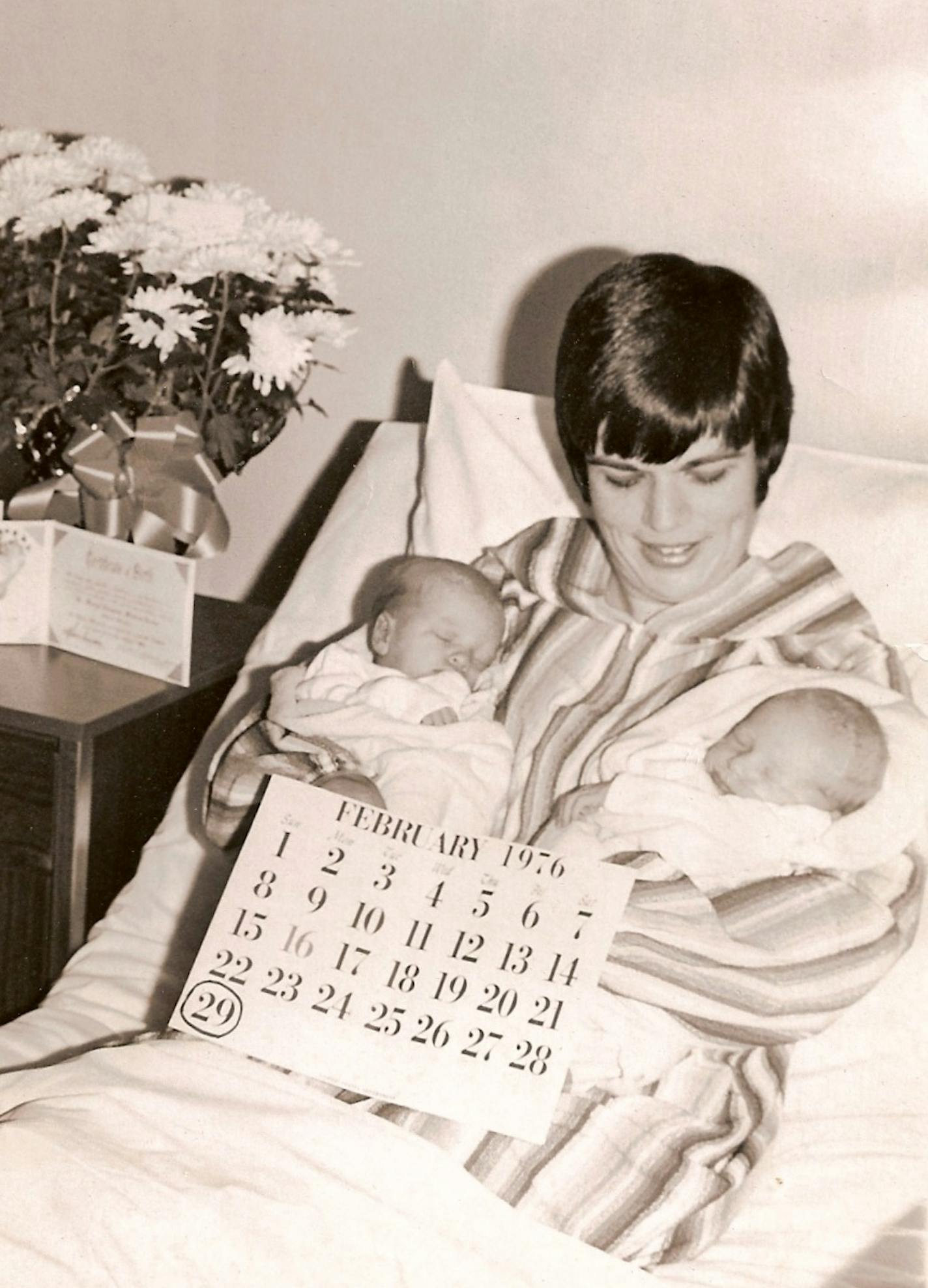 A black and white photo shows a woman with short dark hair smiling while holding two newborn babies in a hospital bed. A calendar of the month of February is displayed in front of them with the 29th circled.