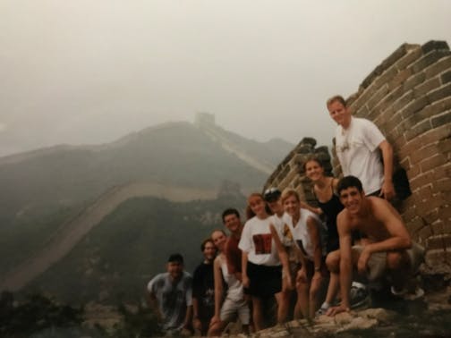 Group of about 10 students and a teacher  looking at the camera along brick facade of the Great Wall of China.