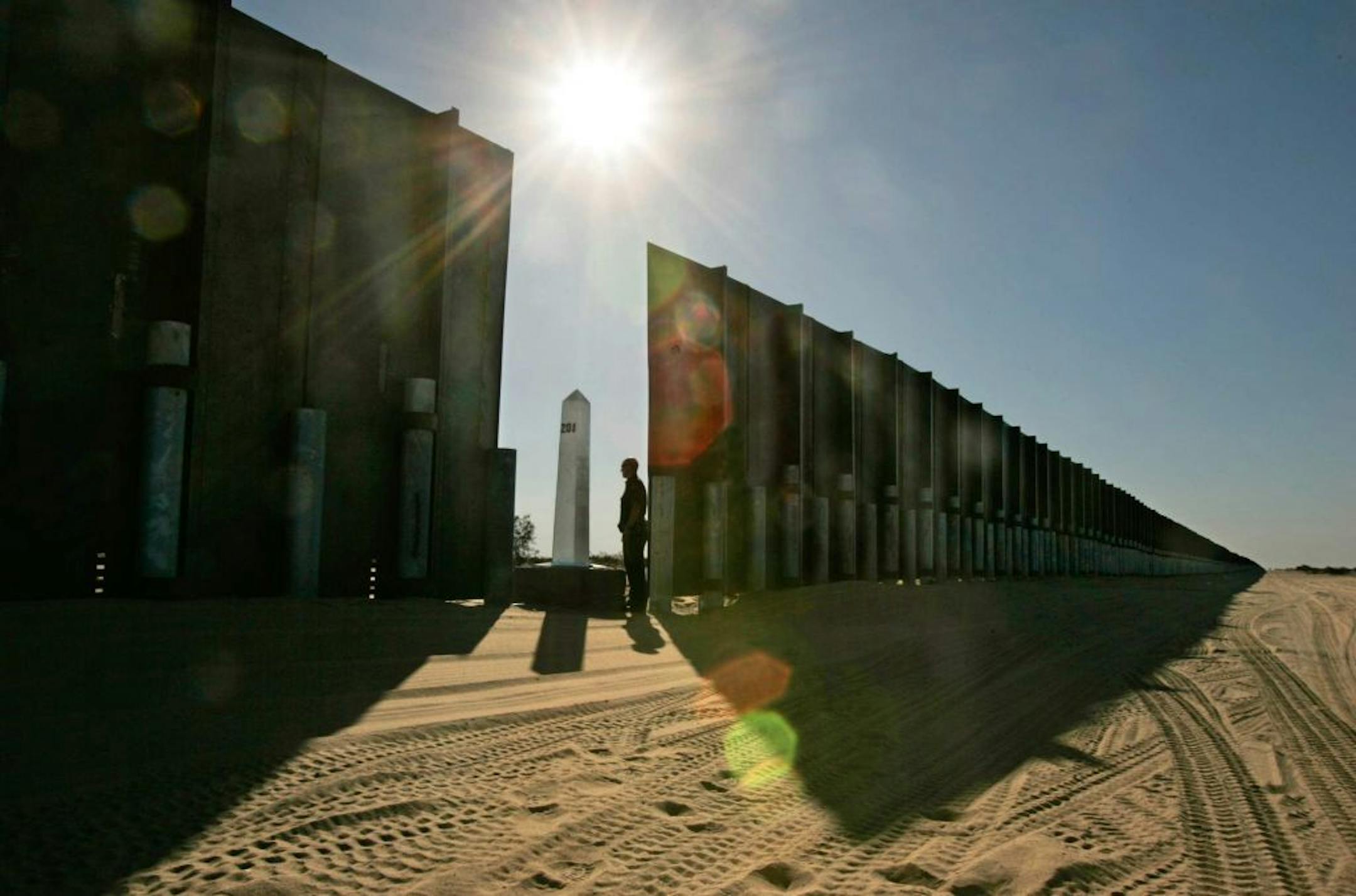 The desert sand east of San Luis, Arizona, is imprinted with border patrol tracks. A gap in the solid steel barrier leaves room for an official international boundary marker.