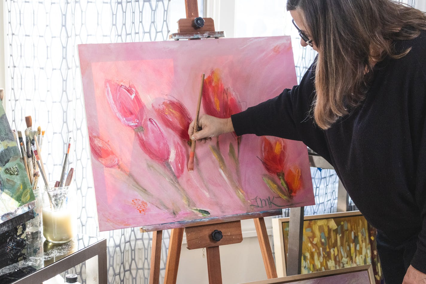 A woman blends brushstrokes onto a canvas upon an easel. The picture shows red and dark pink tulips painted in a contemporary style.