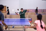 A man attacks a ping pong ball with his paddle while playing against what looks like a father and his daughter. The gym is filled with several table tennis tables, separated by cardboard dividers to keep the balls contained.