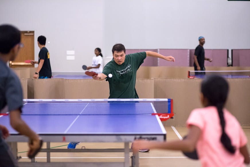 A man attacks a ping pong ball with his paddle while playing against what looks like a father and his daughter. The gym is filled with several table tennis tables, separated by cardboard dividers to keep the balls contained.