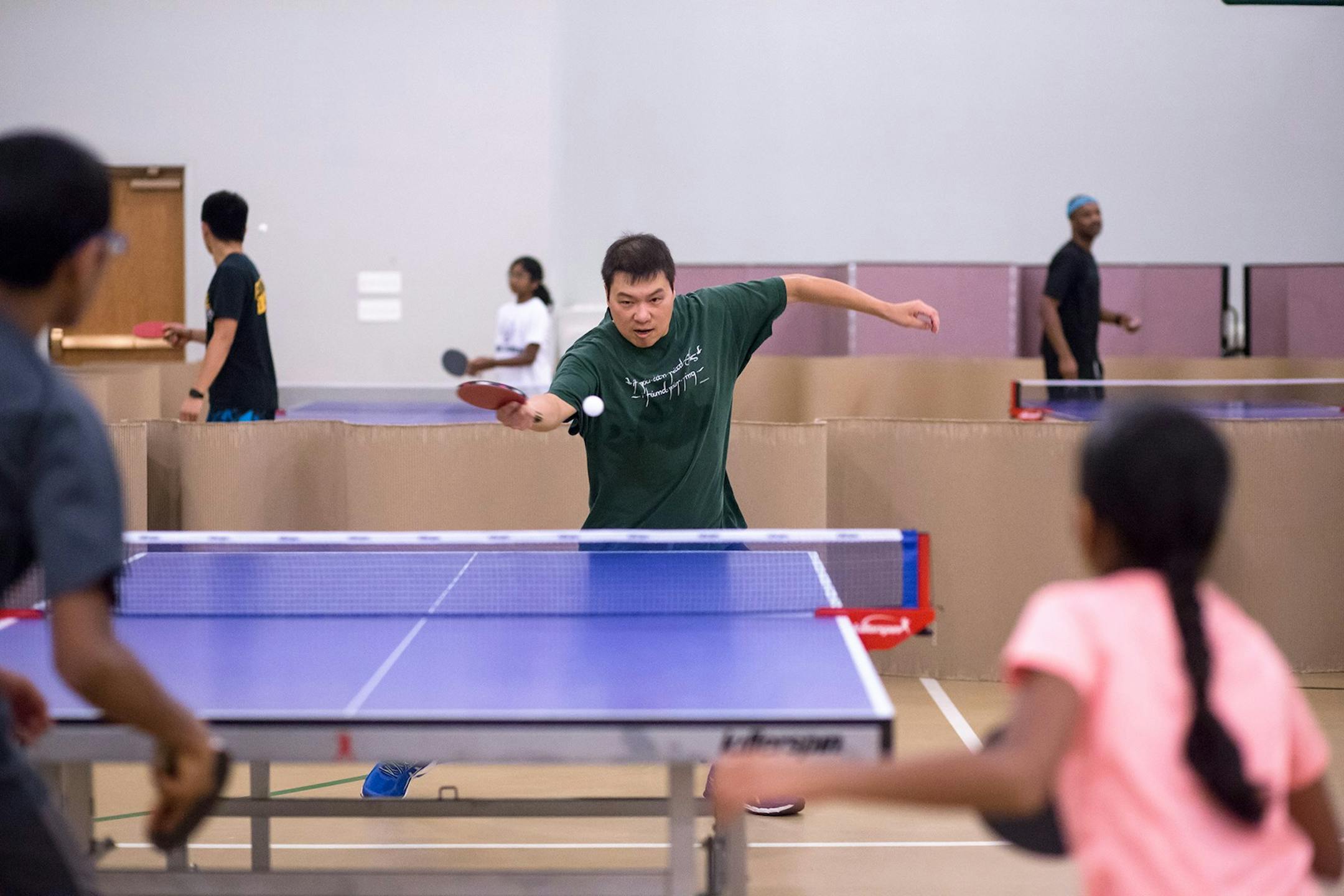 A man attacks a ping pong ball with his paddle while playing against what looks like a father and his daughter. The gym is filled with several table tennis tables, separated by cardboard dividers to keep the balls contained.