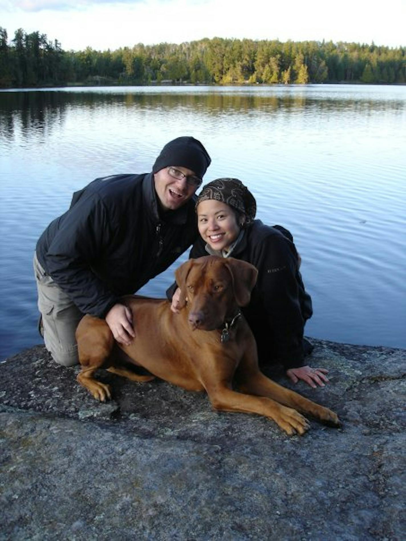 A couple and their large red dog crouch on a rock outcropping with a lake and pine trees in the distance behind them.