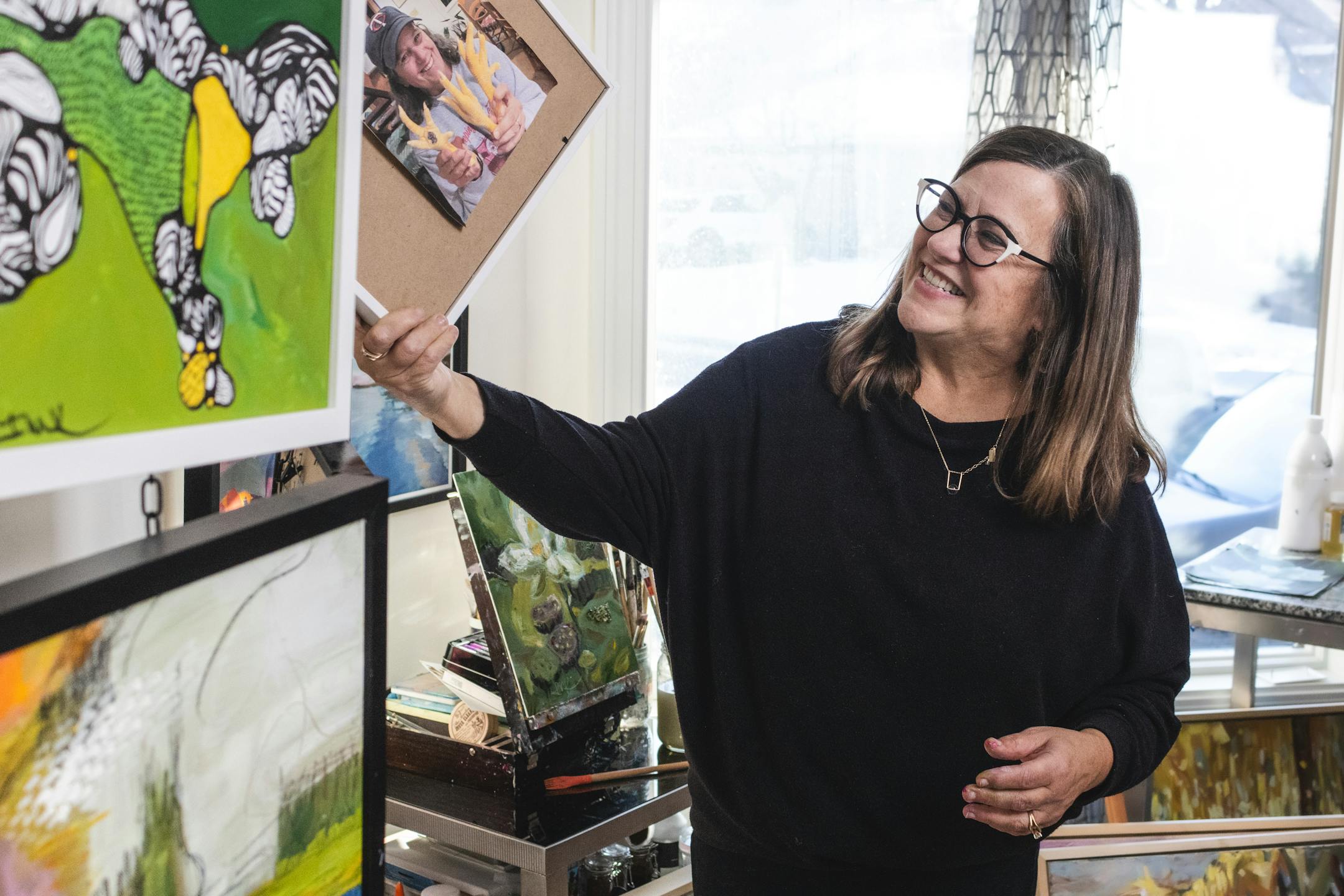 A woman laughs while holding up a framed photograph in a home art studio.