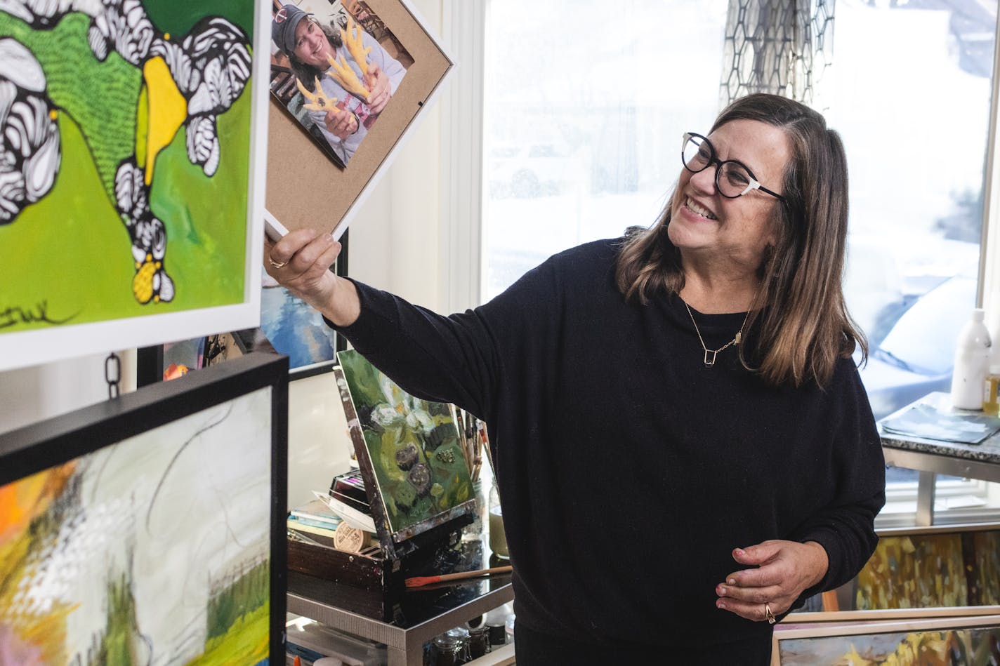 A woman laughs while holding up a framed photograph in a home art studio.